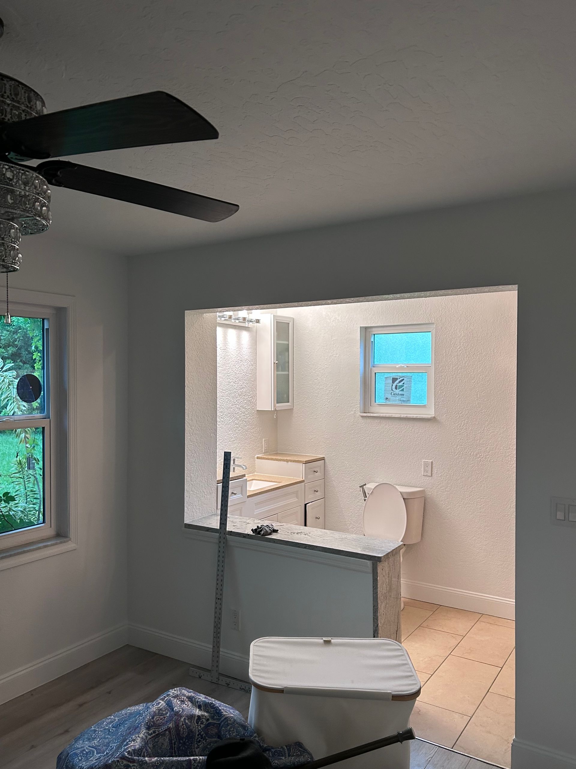 A room with a view into a bathroom and kitchen. Gray walls, ceiling fan, and trash can.