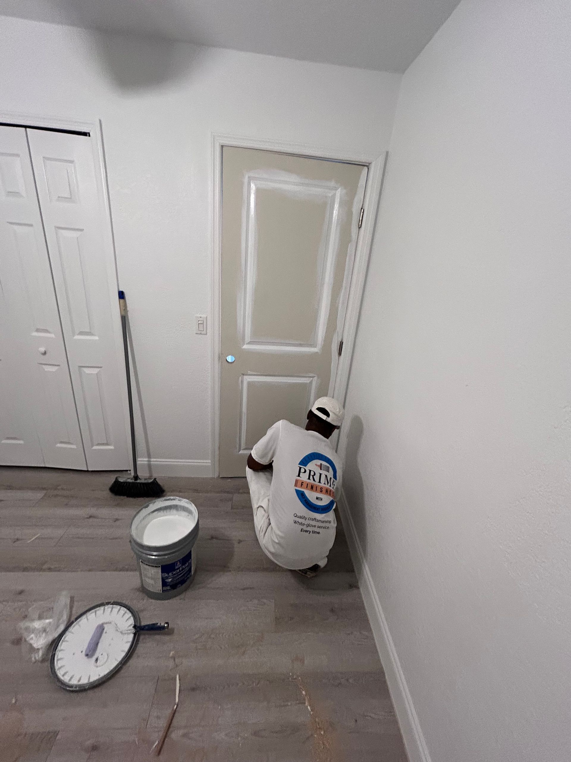 Person painting a door in a room with white walls and gray flooring; paint bucket and scale on the floor.