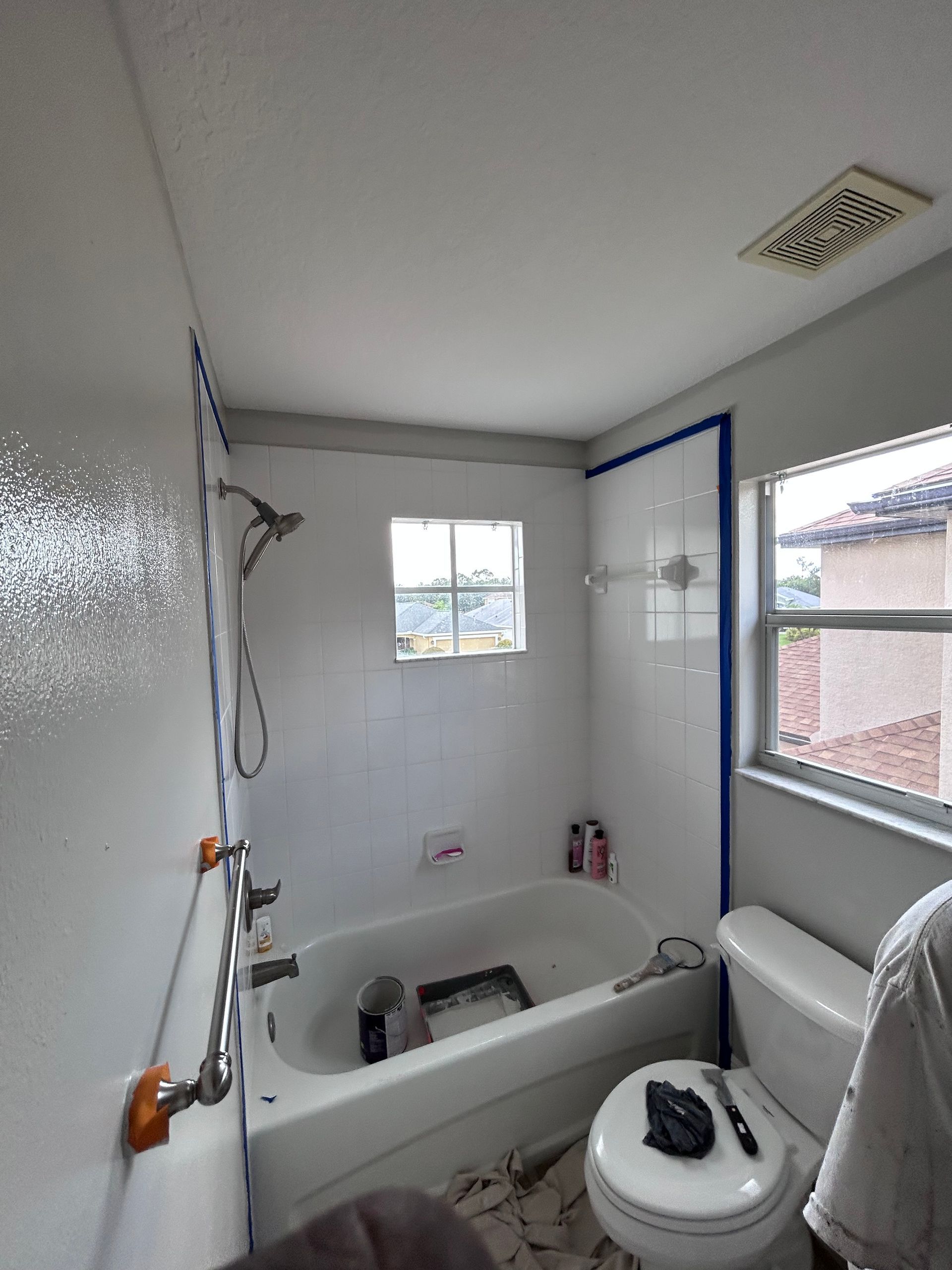 Bathroom undergoing renovation with blue painter's tape, bathtub, toilet, window, and paint supplies.
