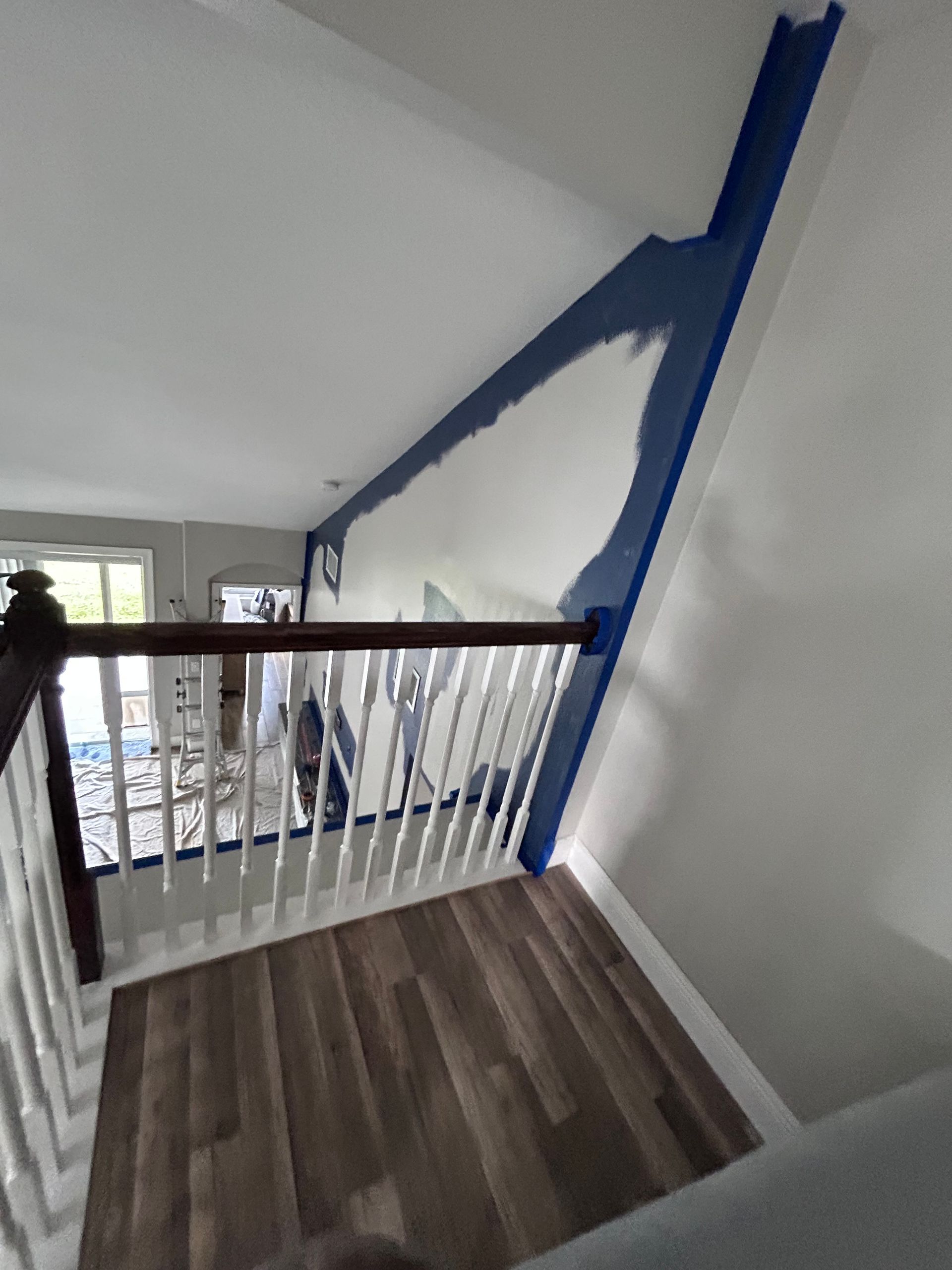 A partially painted blue accent wall with tape, above a staircase with a white railing and wooden floors.