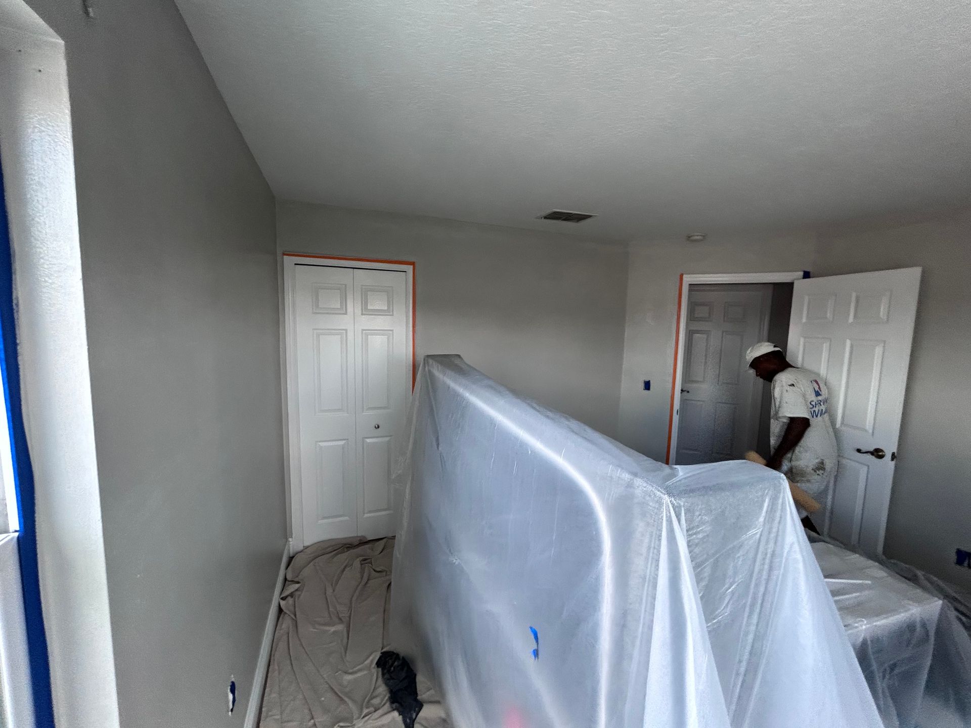 Room interior being painted; painter covers furniture with plastic, walls are grey, doors are white.
