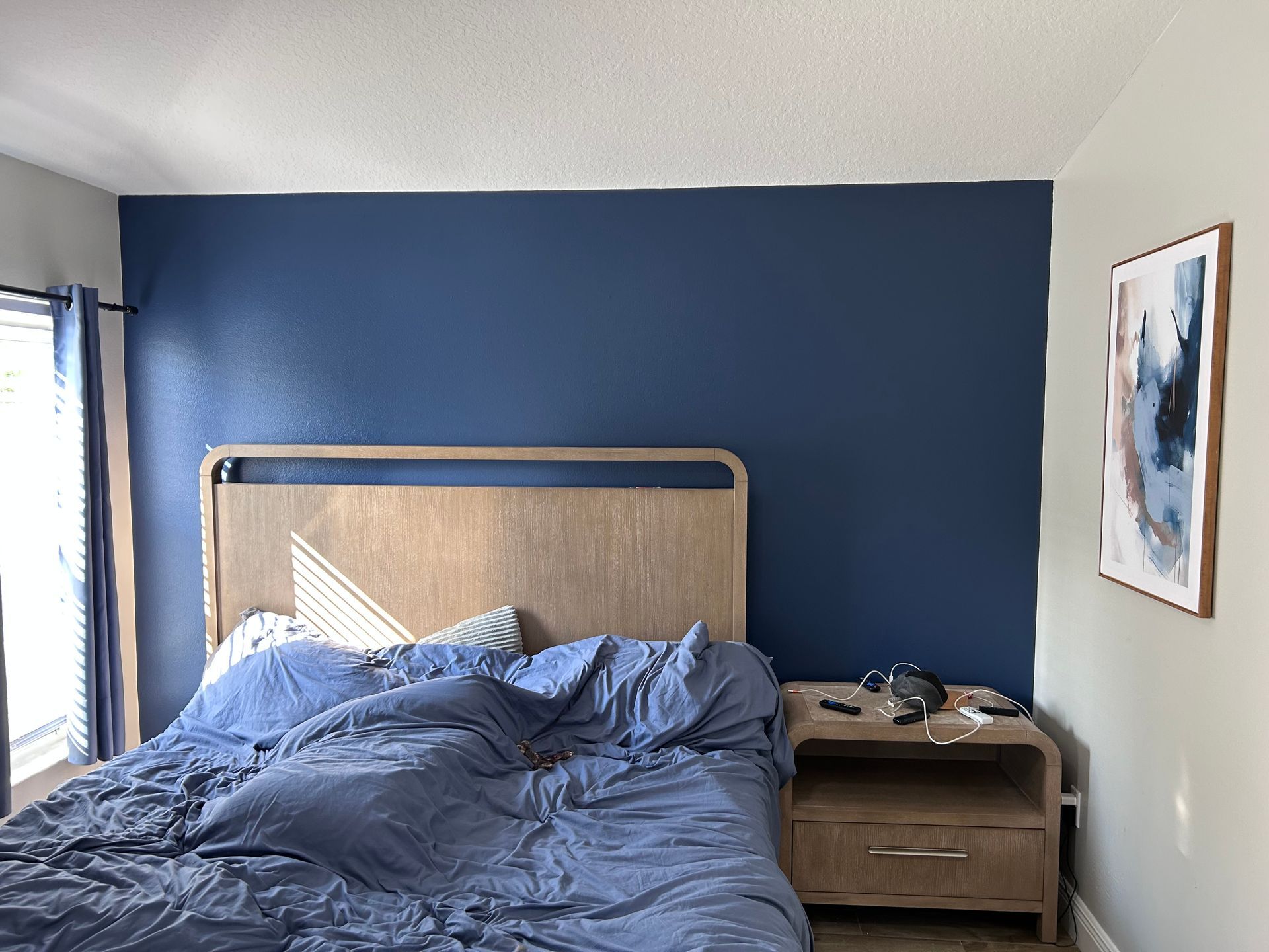 Bedroom with unmade bed, blue wall, wooden nightstand, and abstract art.