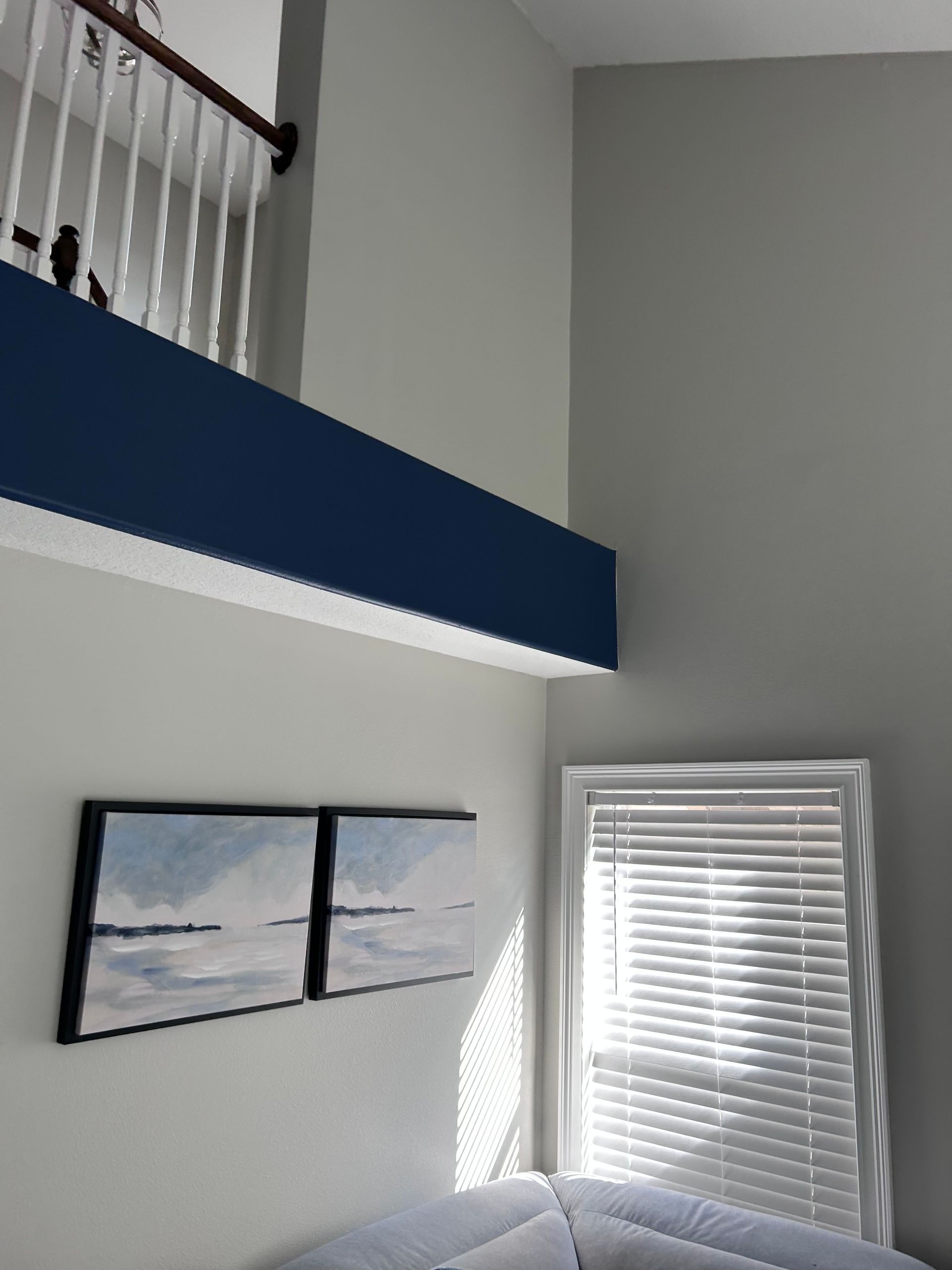 A room with a high ceiling, a blue beam, a window with blinds, and artwork. Sunlight is streaming in.