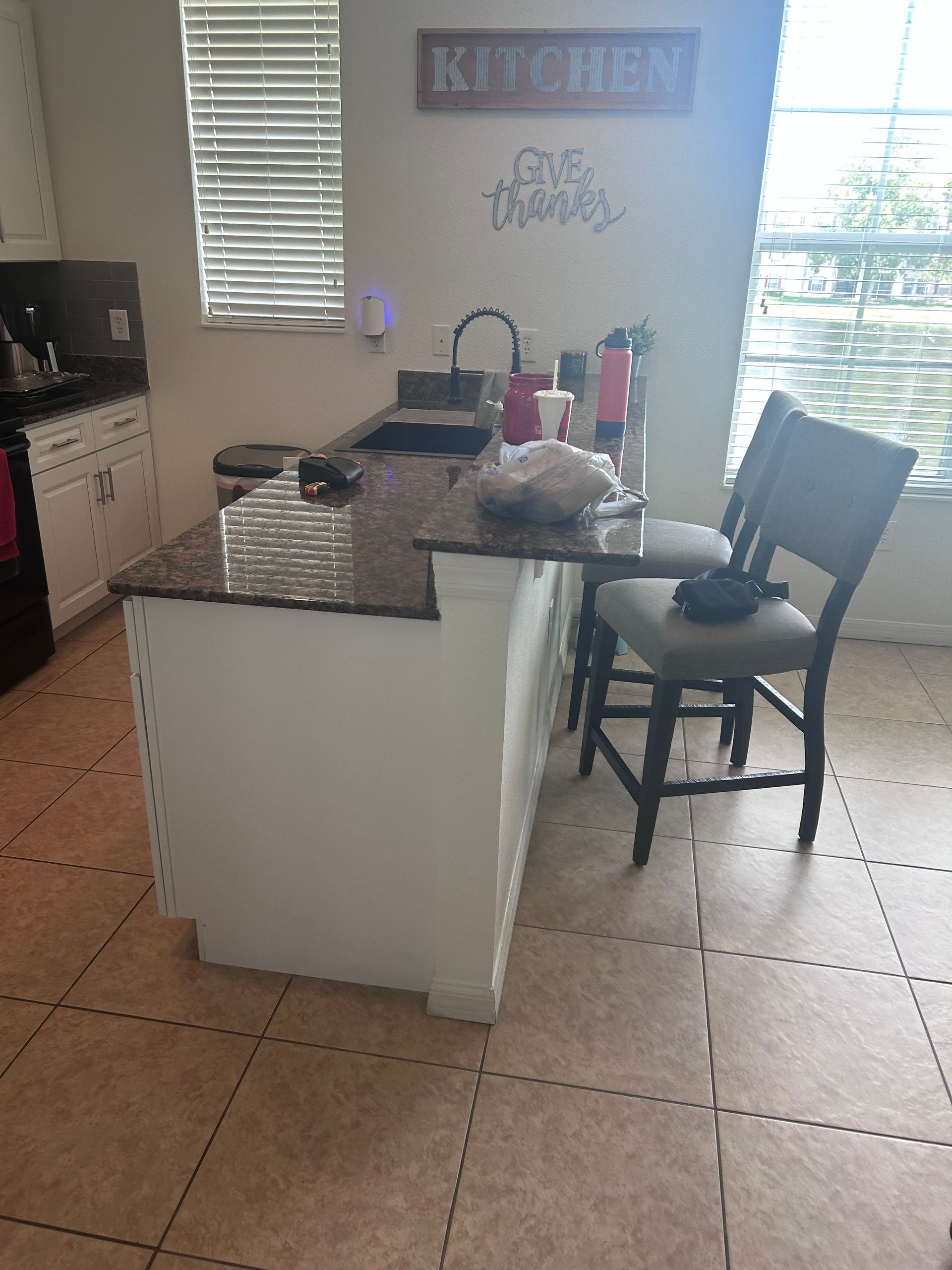 Kitchen with island, black countertop, white cabinets, brown tile floor, and a chair.