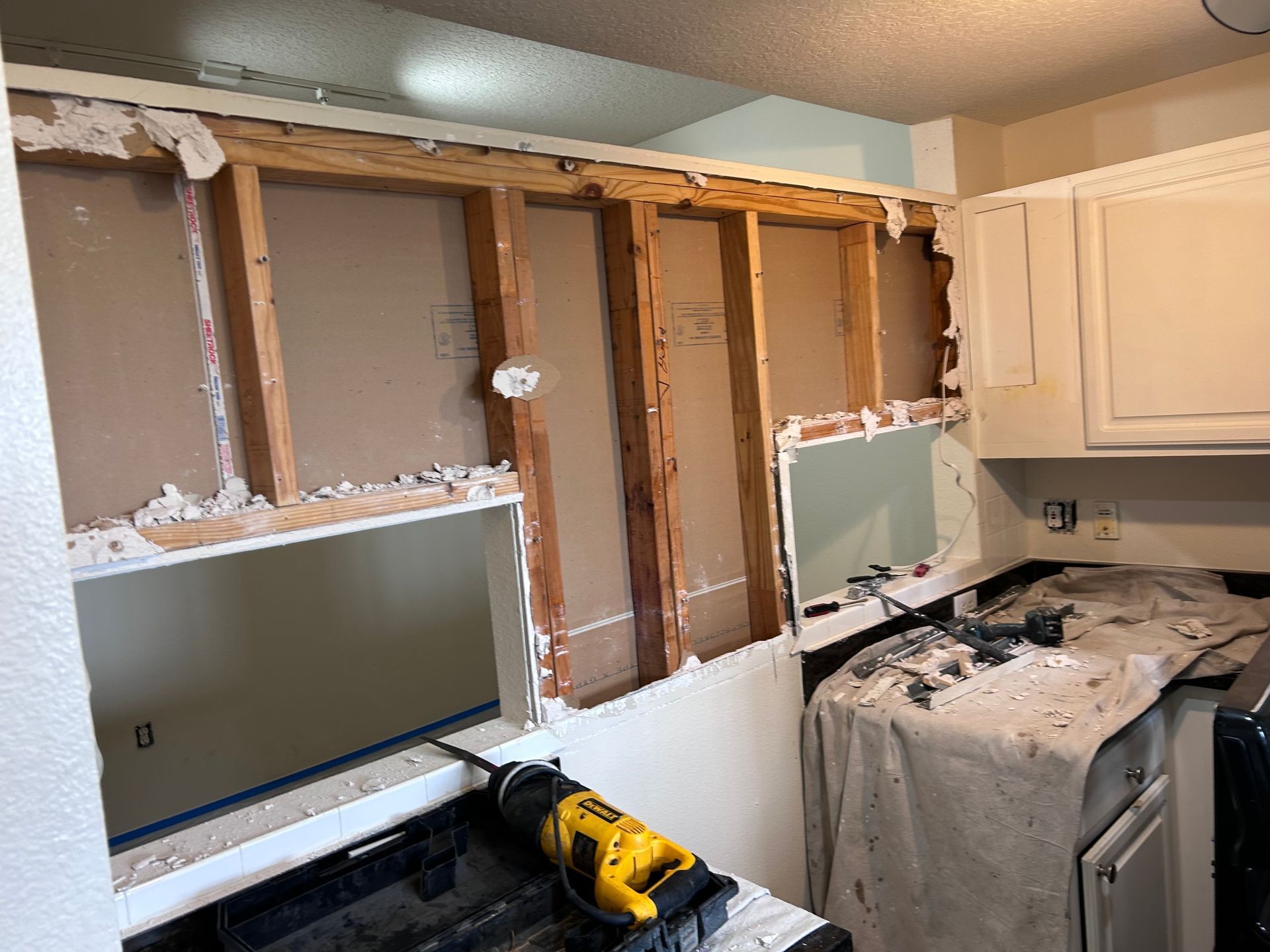 Kitchen wall demolition in progress, exposing studs and drywall removal. Yellow drill on counter.