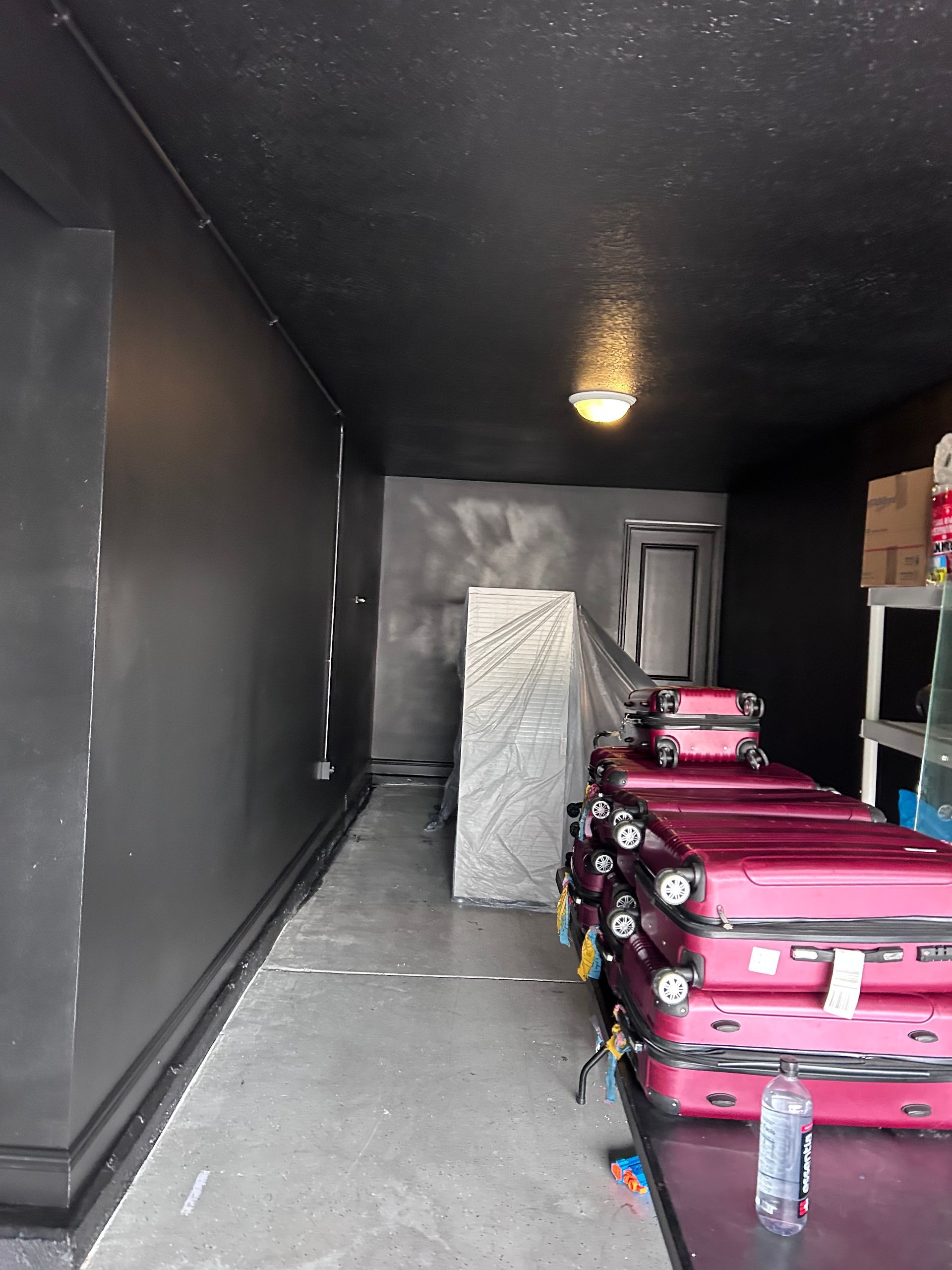 Narrow storage room with a stack of pink suitcases, a stone column, and black walls.