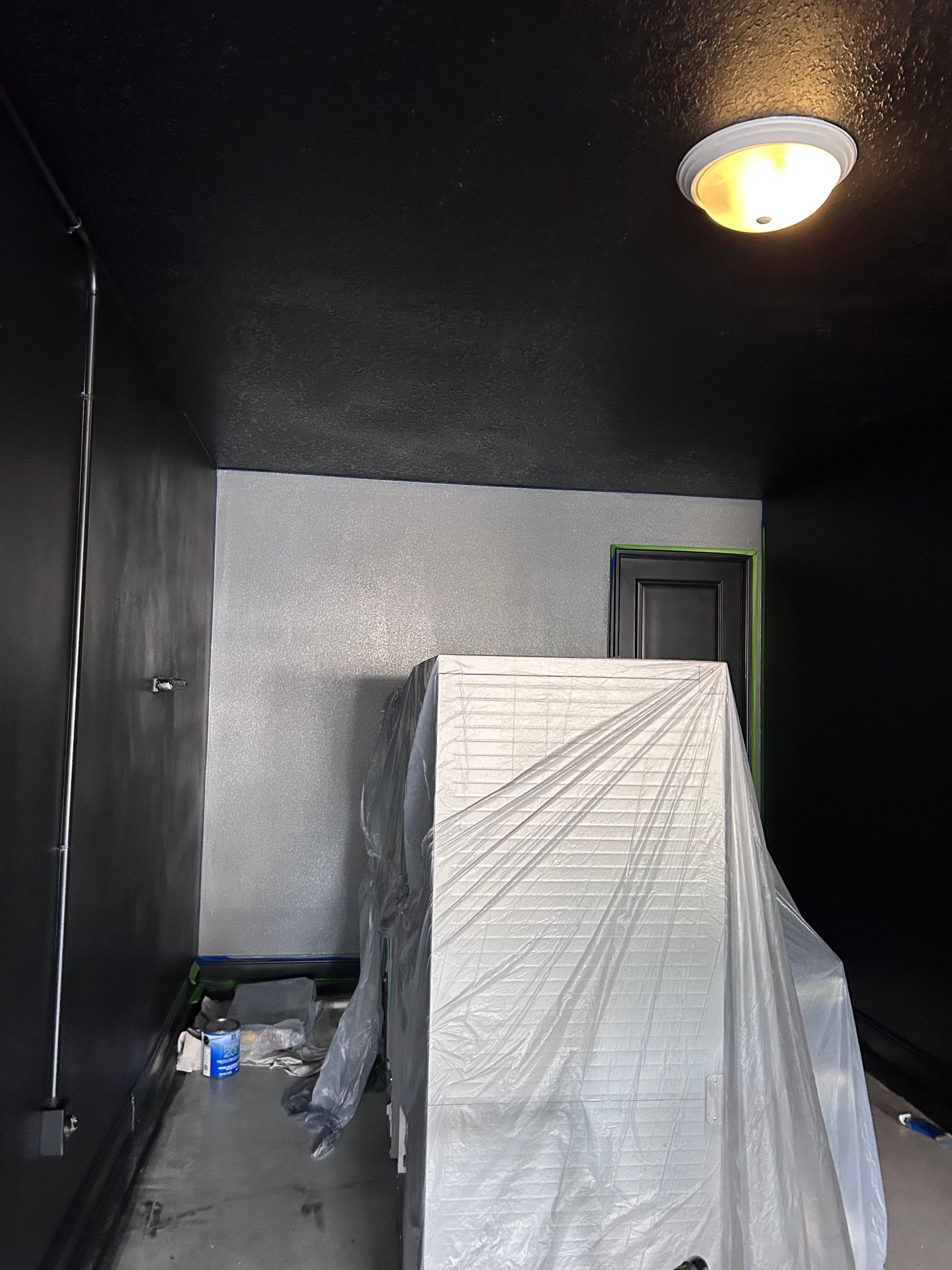Room with black walls and ceiling, silver textured wall. Object covered in plastic. Ceiling light.