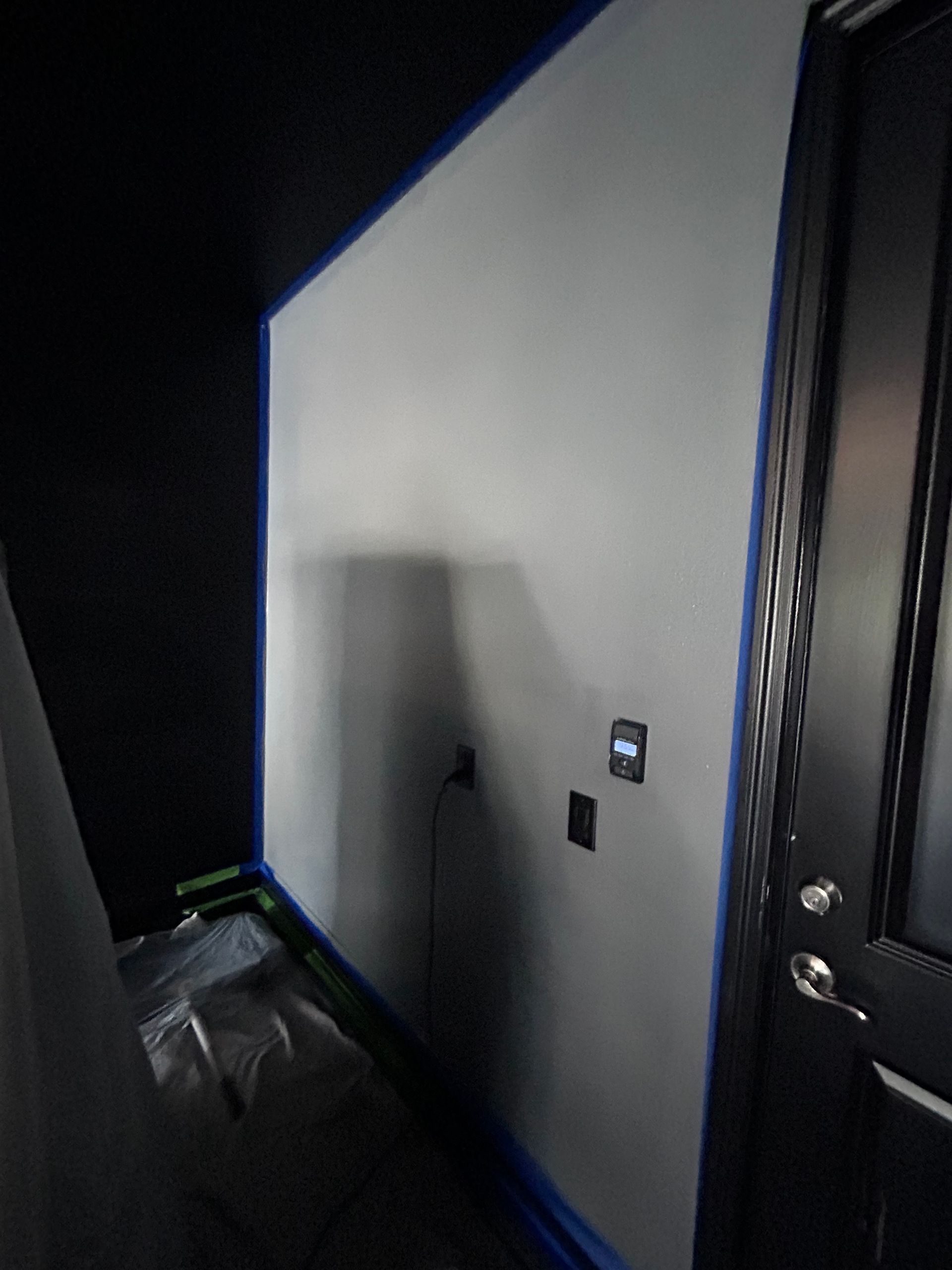 Interior wall being painted white with blue tape, next to a black wall and door.