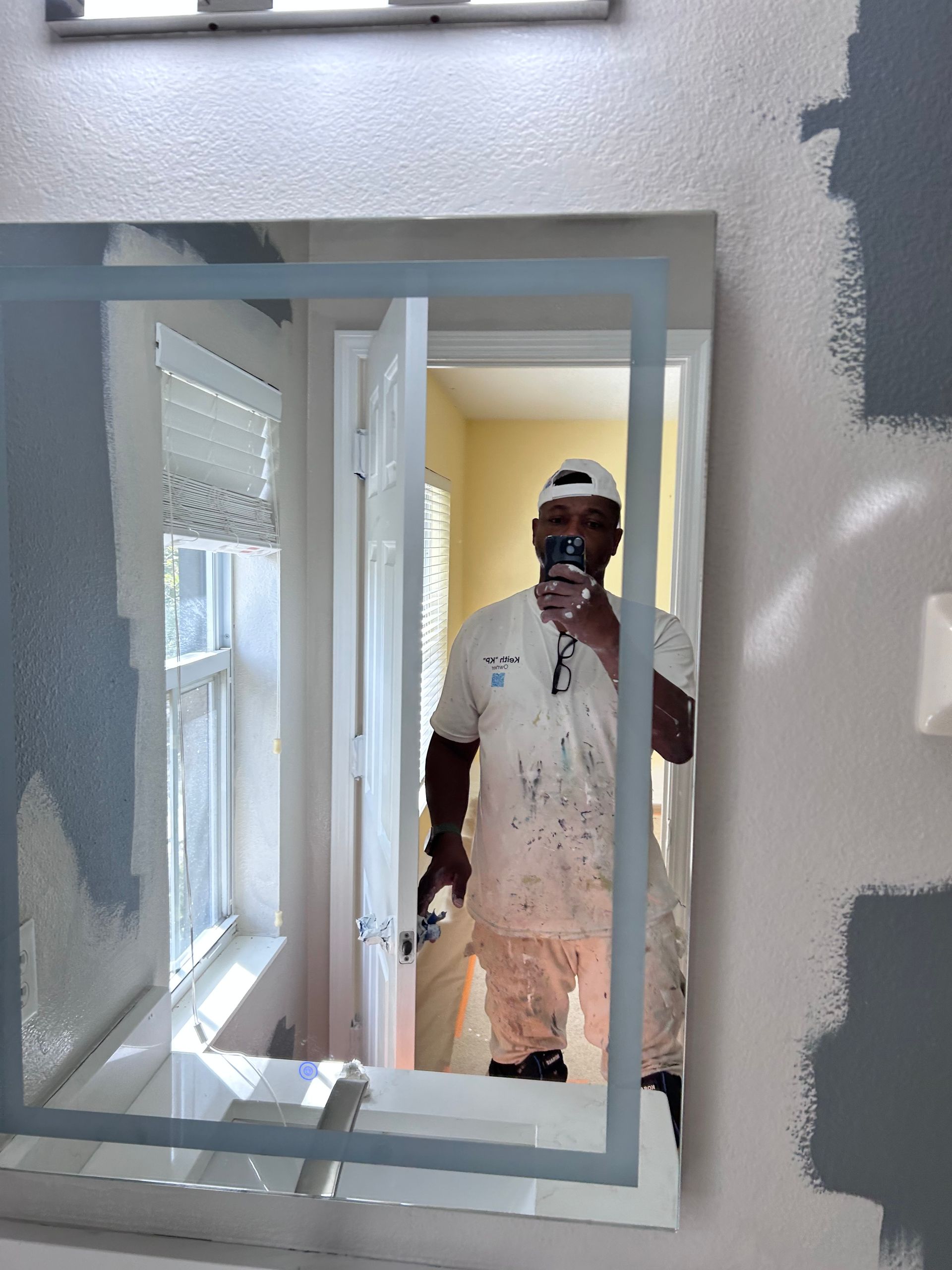 Man in paint-splattered clothes taking a mirror selfie. He’s in a bathroom with gray paint swatches on the wall.