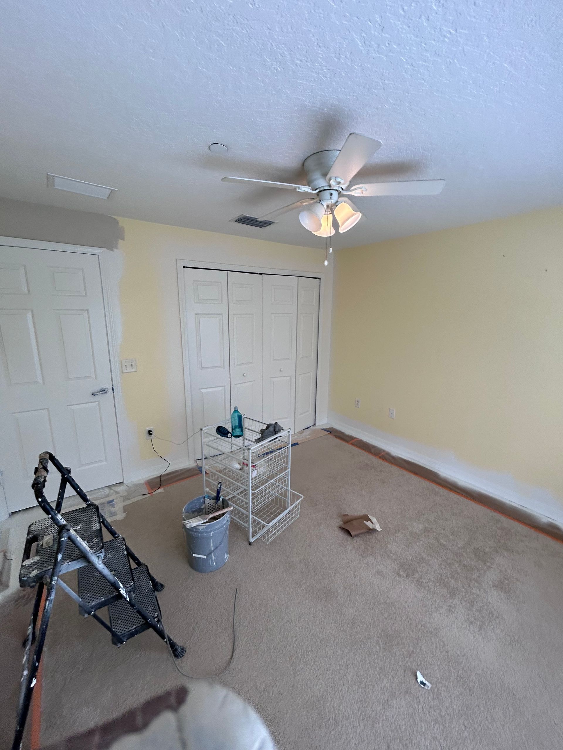 Room with yellow walls, white closet doors, ceiling fan, and ladder; carpeted floor.
