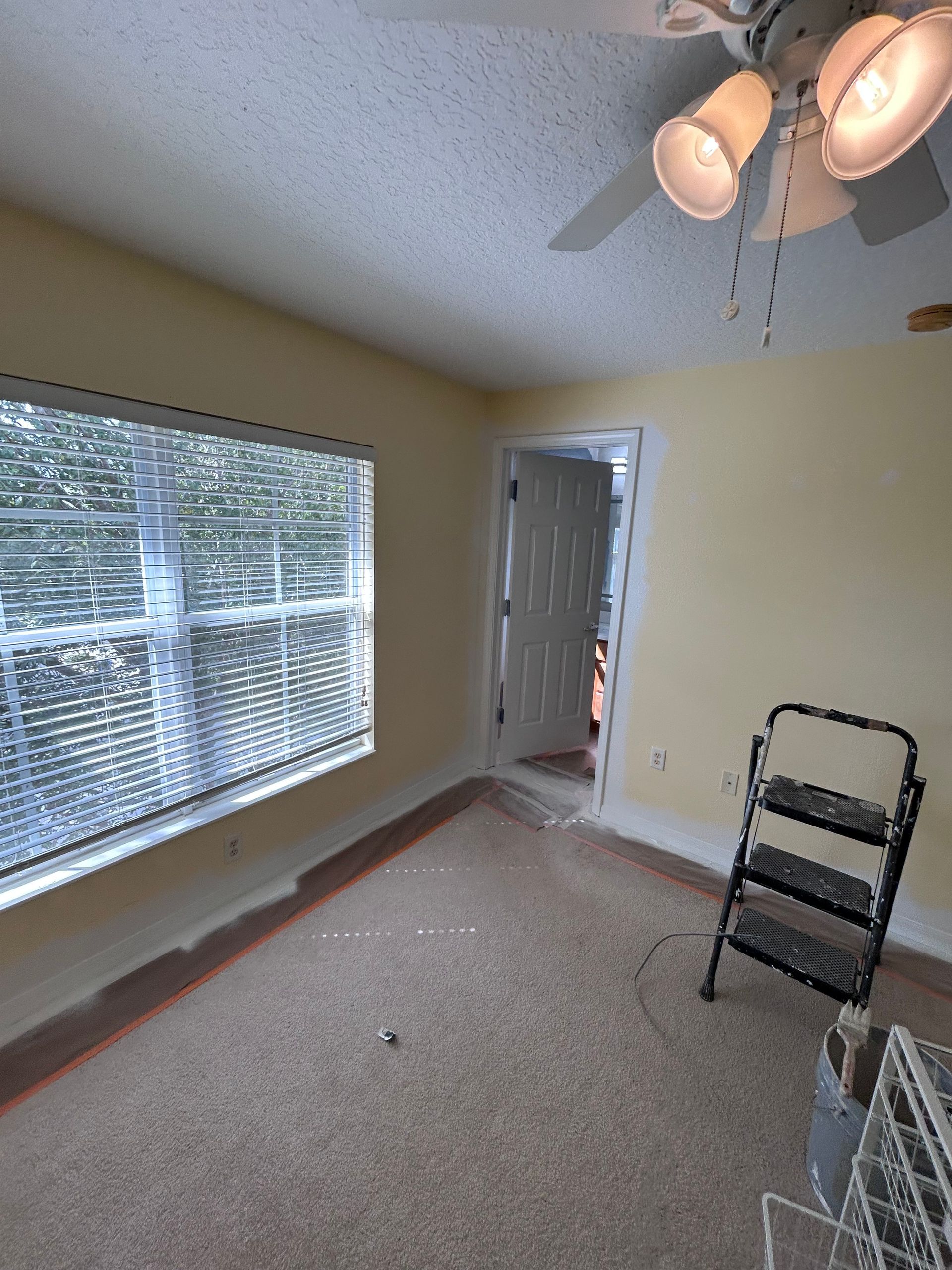 Empty room with yellow walls, window with blinds, open door, ceiling fan, and step ladder.