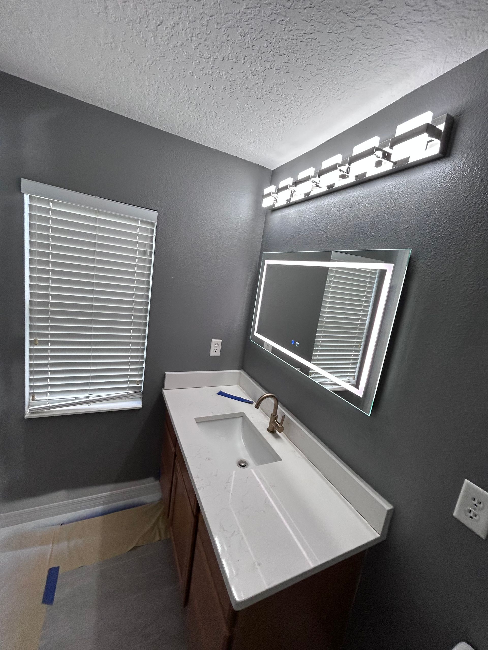 Bathroom with dark gray walls, white countertop, lighted mirror, and blinds.