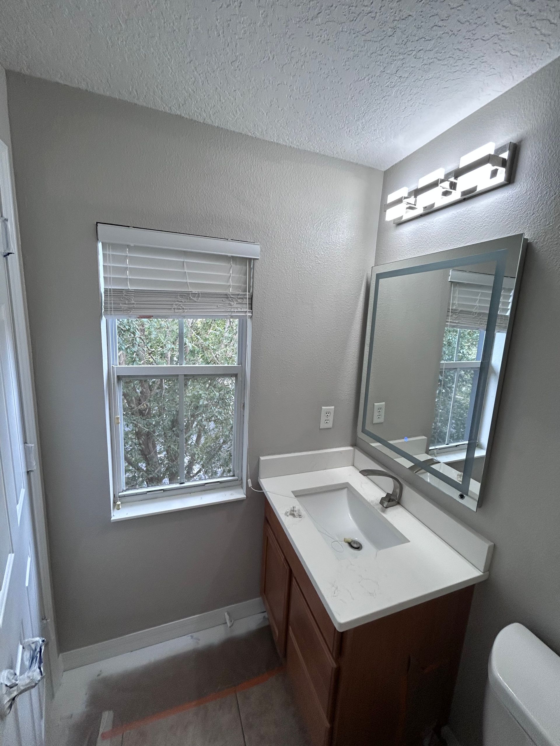 Bathroom with a vanity, window, mirror, and toilet. Beige walls, wood cabinet, and white countertop.