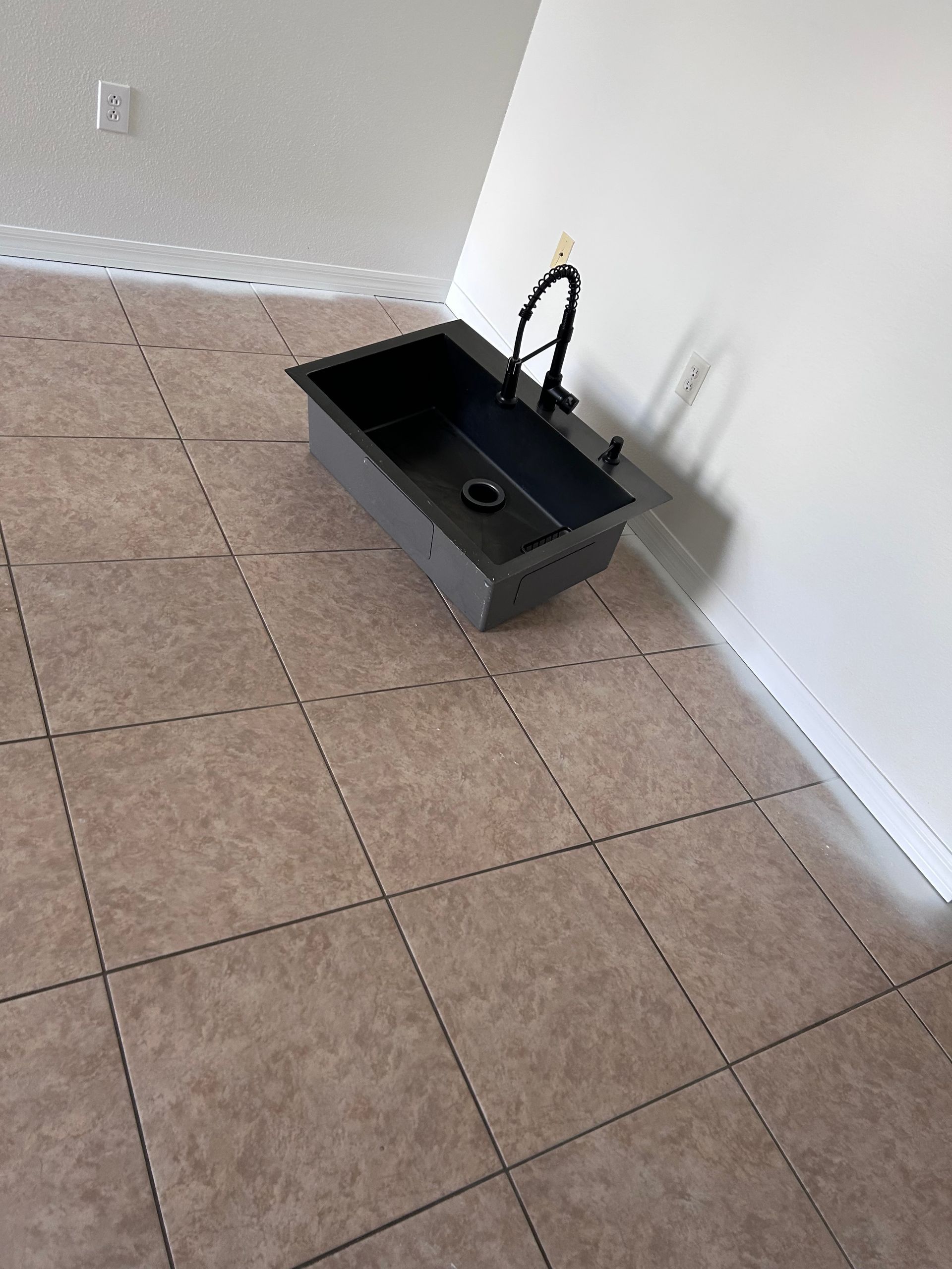 Dark sink and faucet on floor, near a white wall. Brown tile floor.