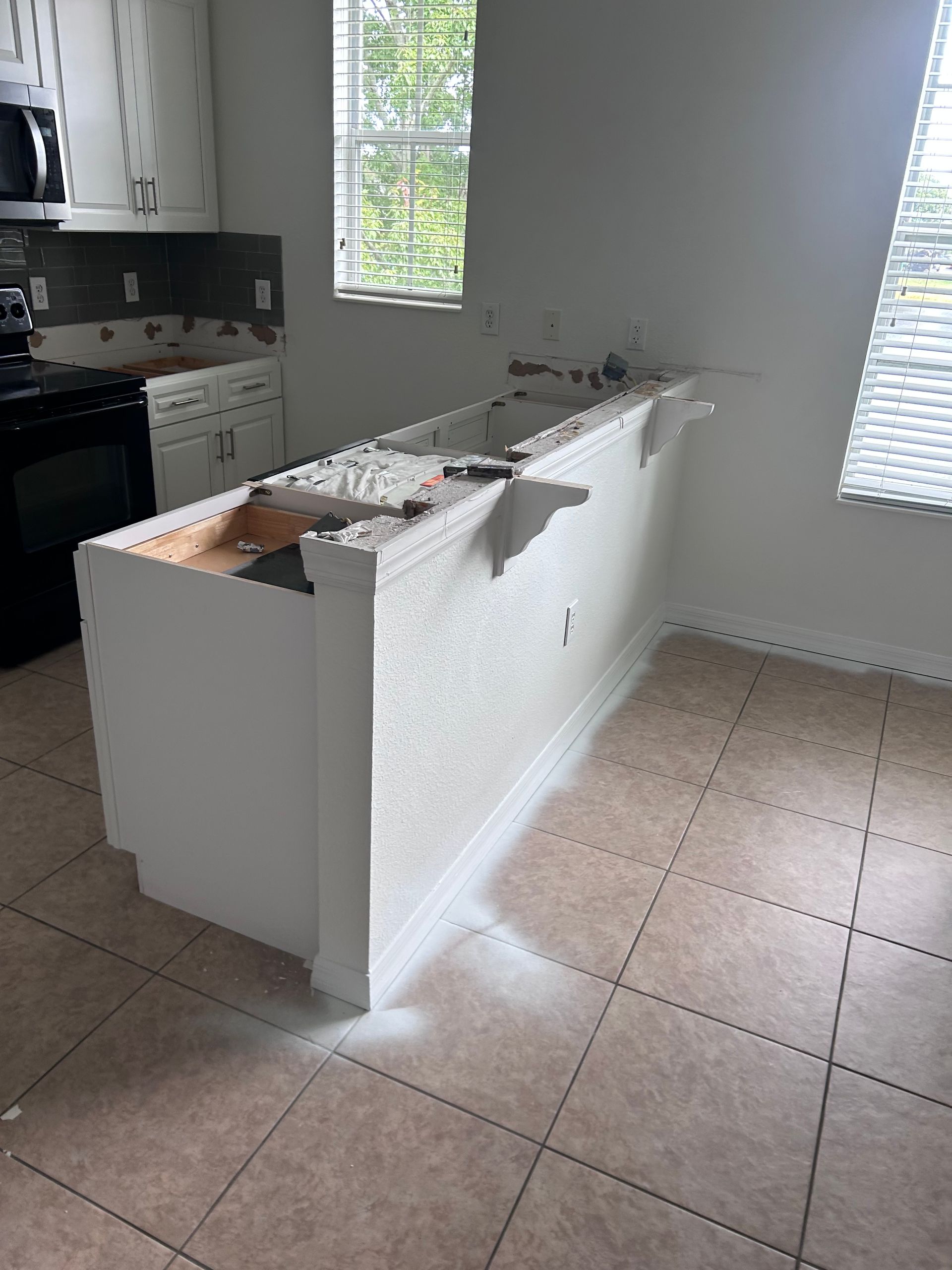 Kitchen island under renovation, missing countertop. White cabinets, black stove, tiled floor. Sunlight through window.