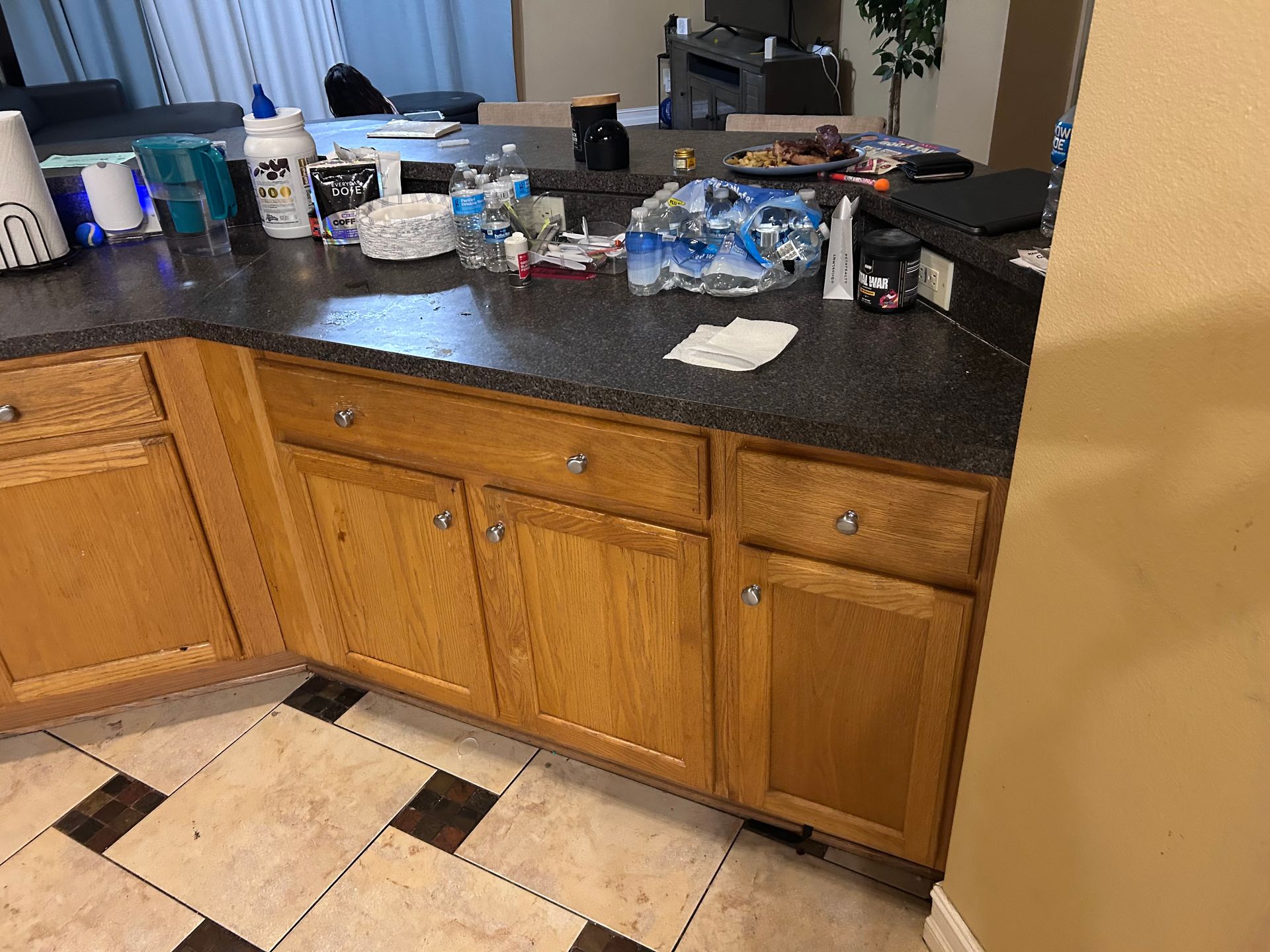Kitchen counter with light wood cabinets, dark countertop, and various items scattered across the surface.