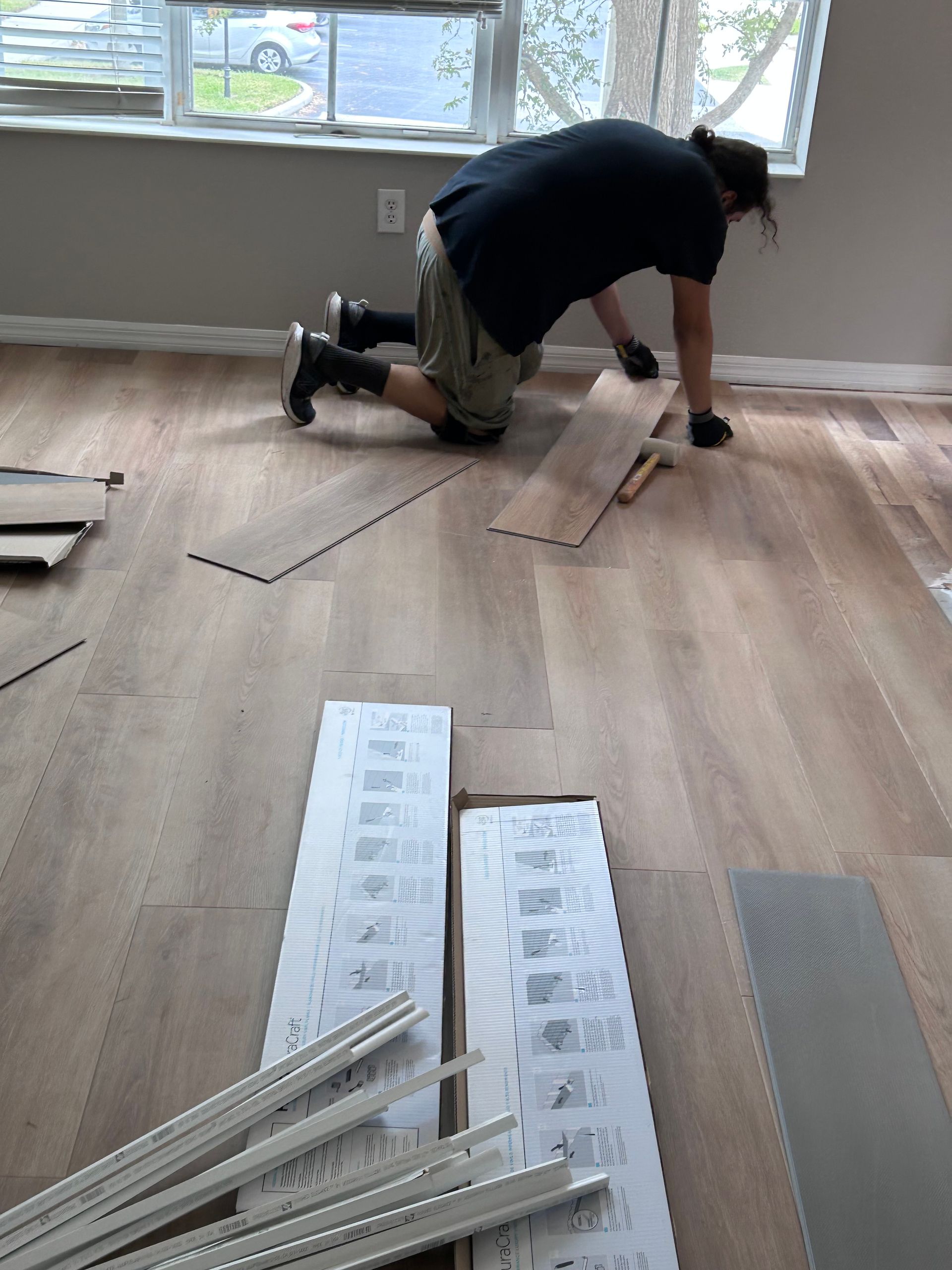 Person kneeling, installing wood flooring in a room with a window, boxes of planks and molding nearby.