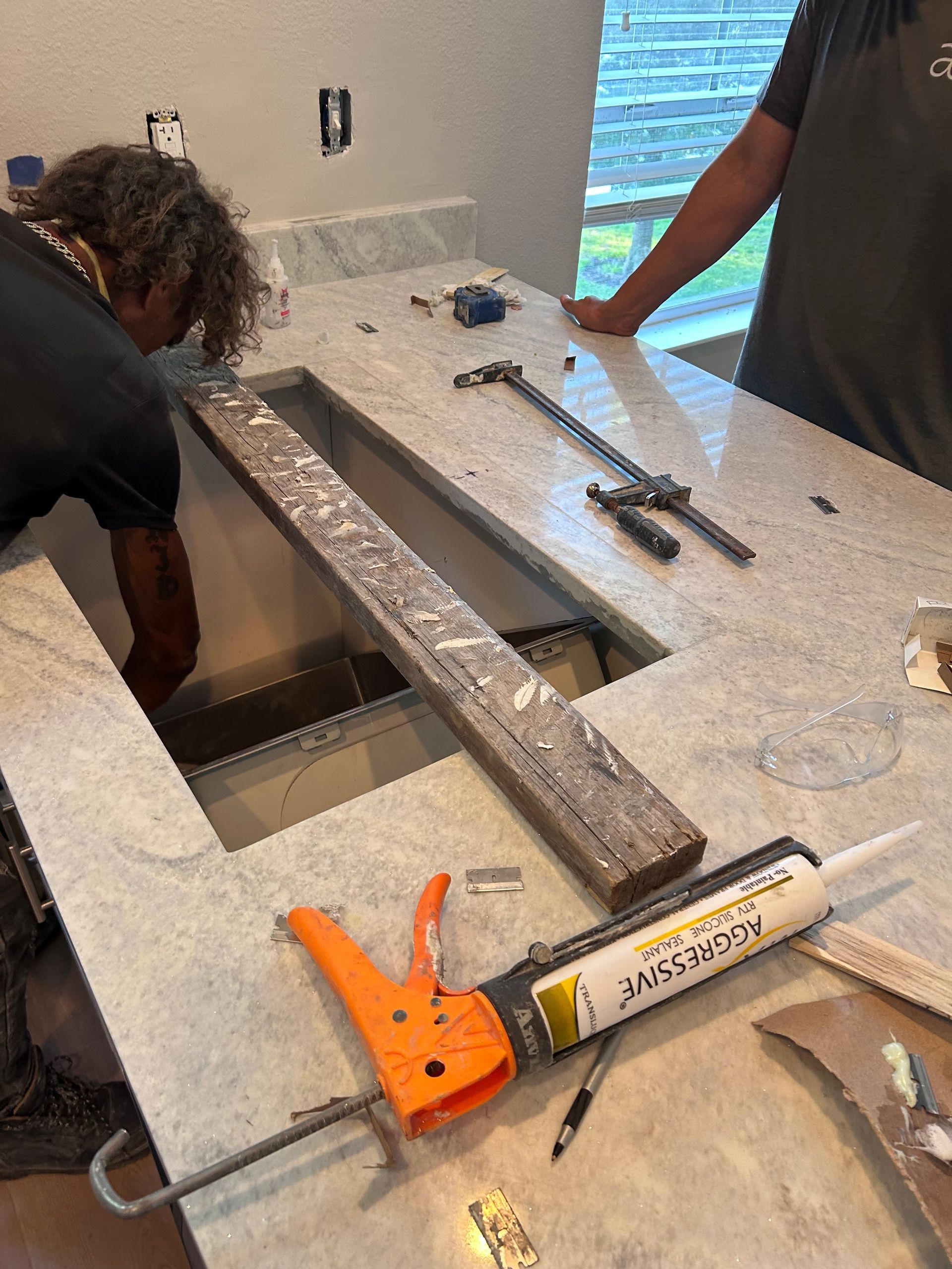 Two people installing a countertop with a sink cutout. Adhesive gun and tools on the granite surface.