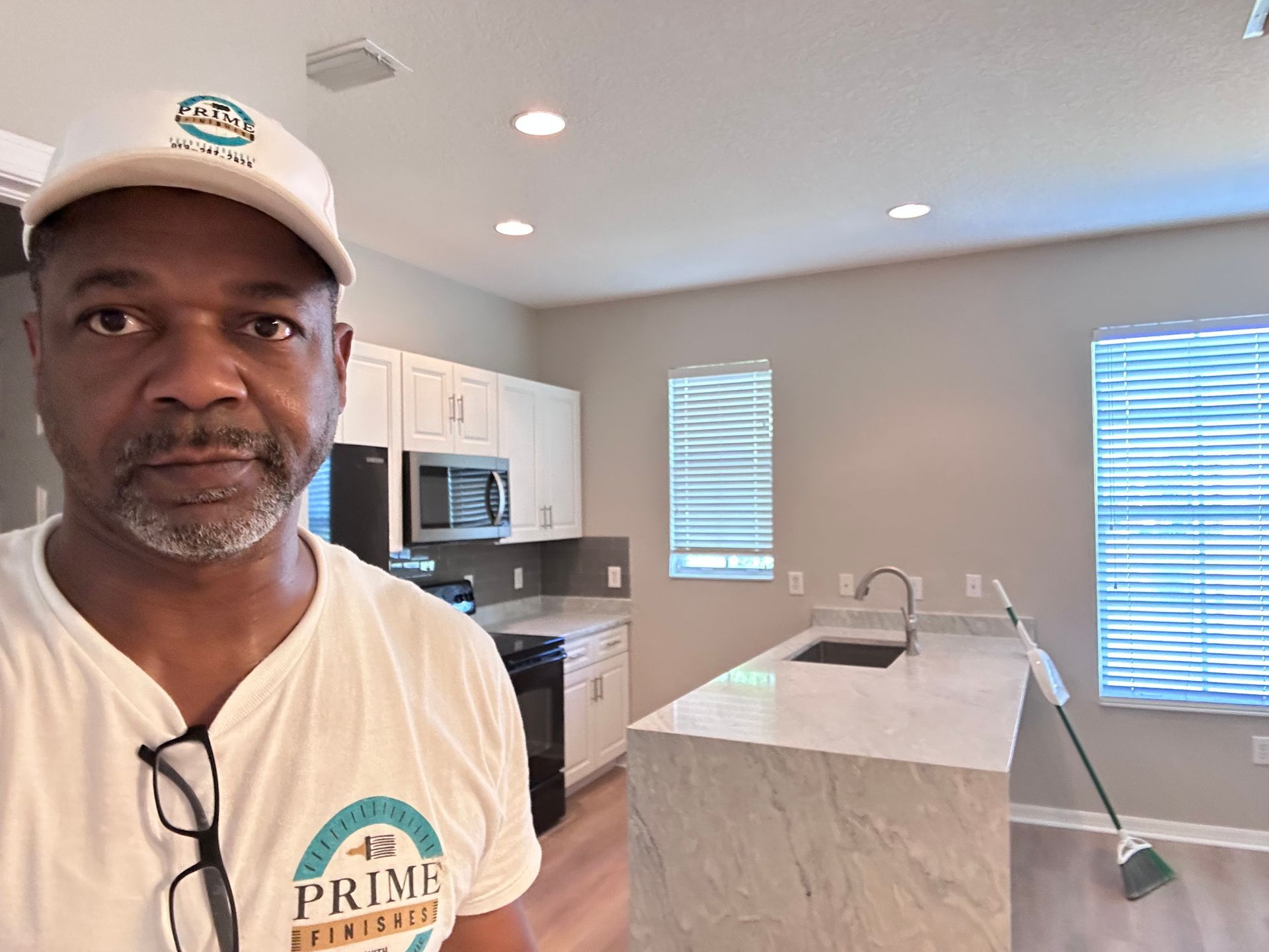 Man in white hat and shirt stands in a modern kitchen. White cabinets, marble countertop, and windows.