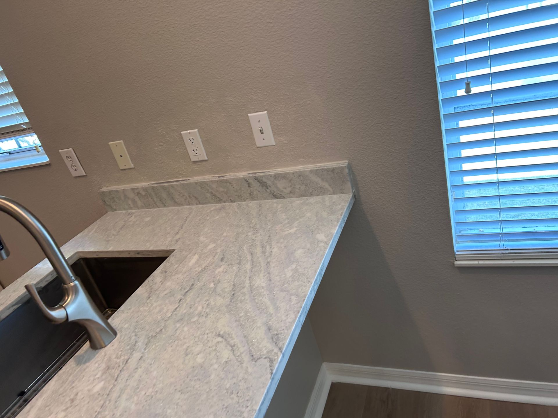 Kitchen countertop with sink and faucet, outlets, and window with blinds.