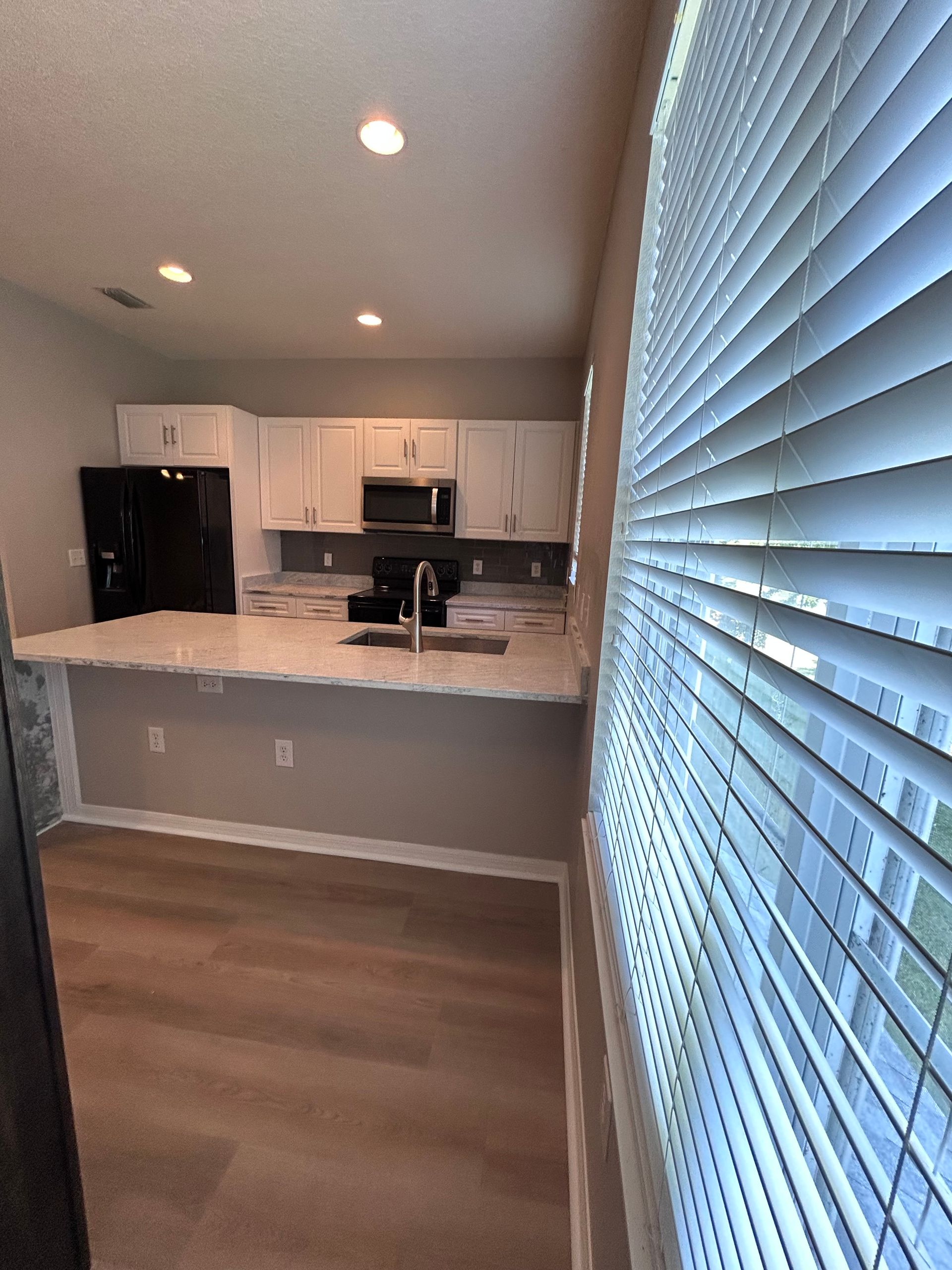 Kitchen with white cabinets, countertops, and black appliances. Blinds partially cover a window.