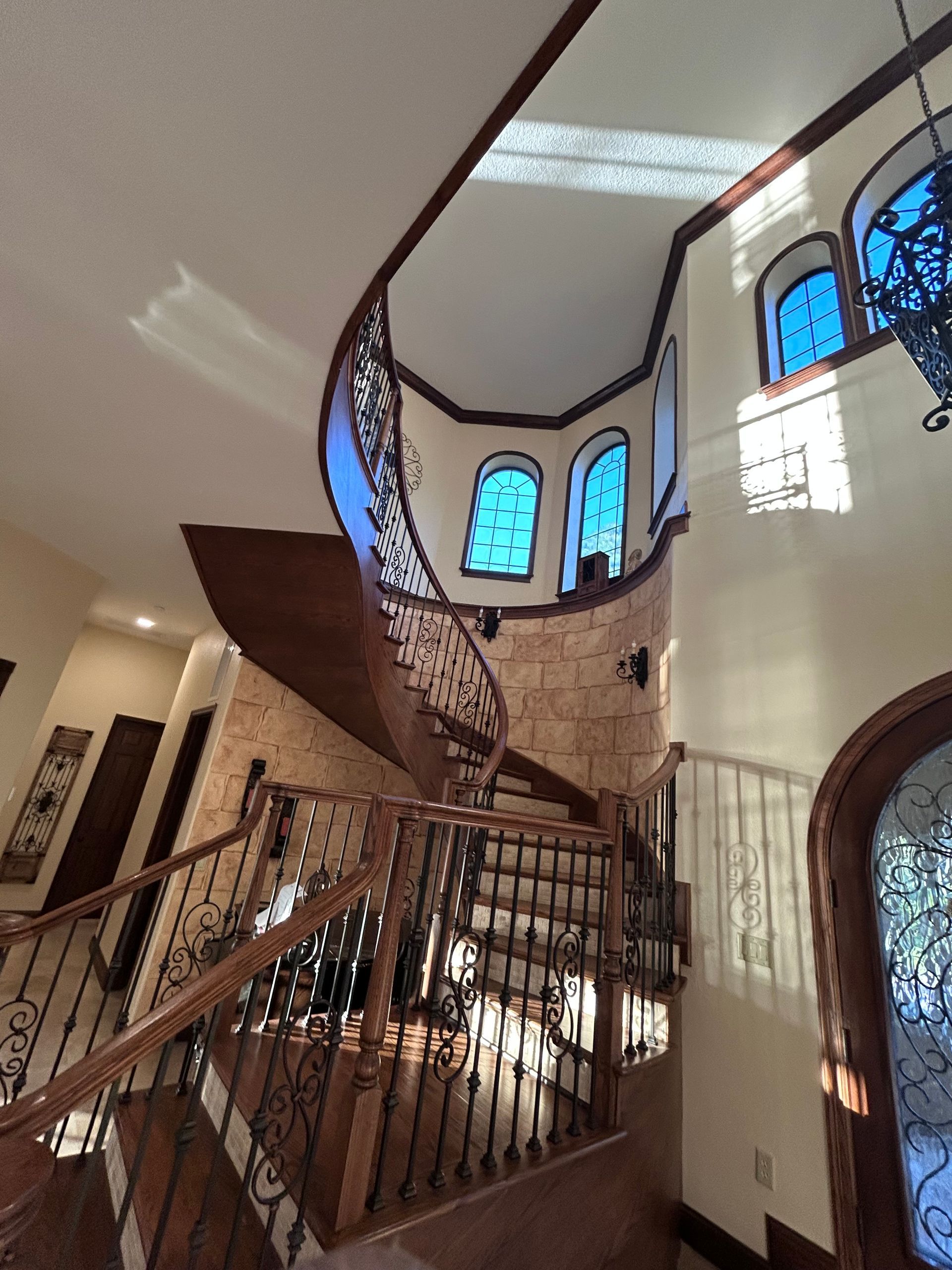 Ornate, curving staircase with dark wood and wrought iron details against a textured stone wall. Blue stained-glass windows are above.