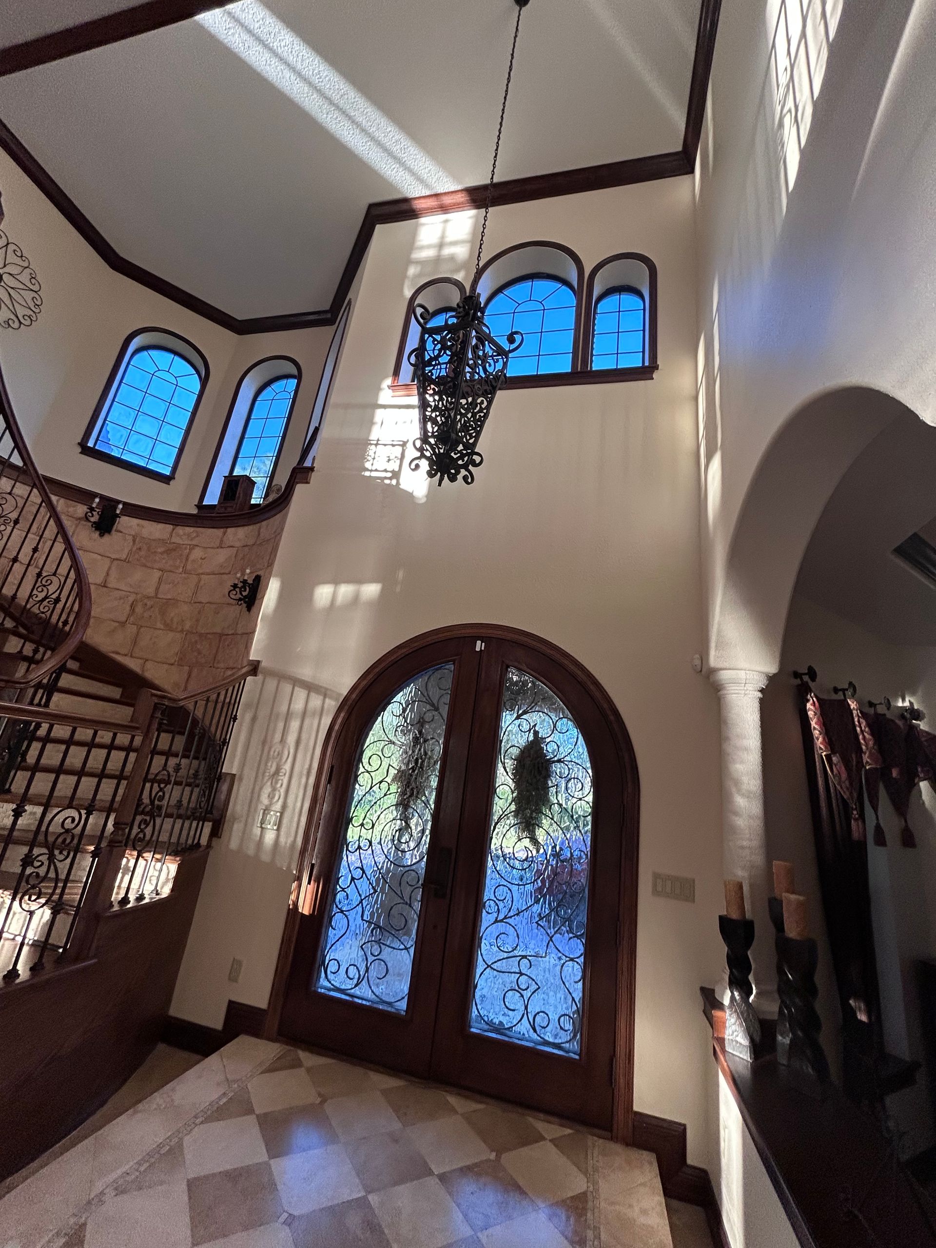 Foyer with arched wooden doors, a winding staircase, blue-tinted windows, and a hanging chandelier.