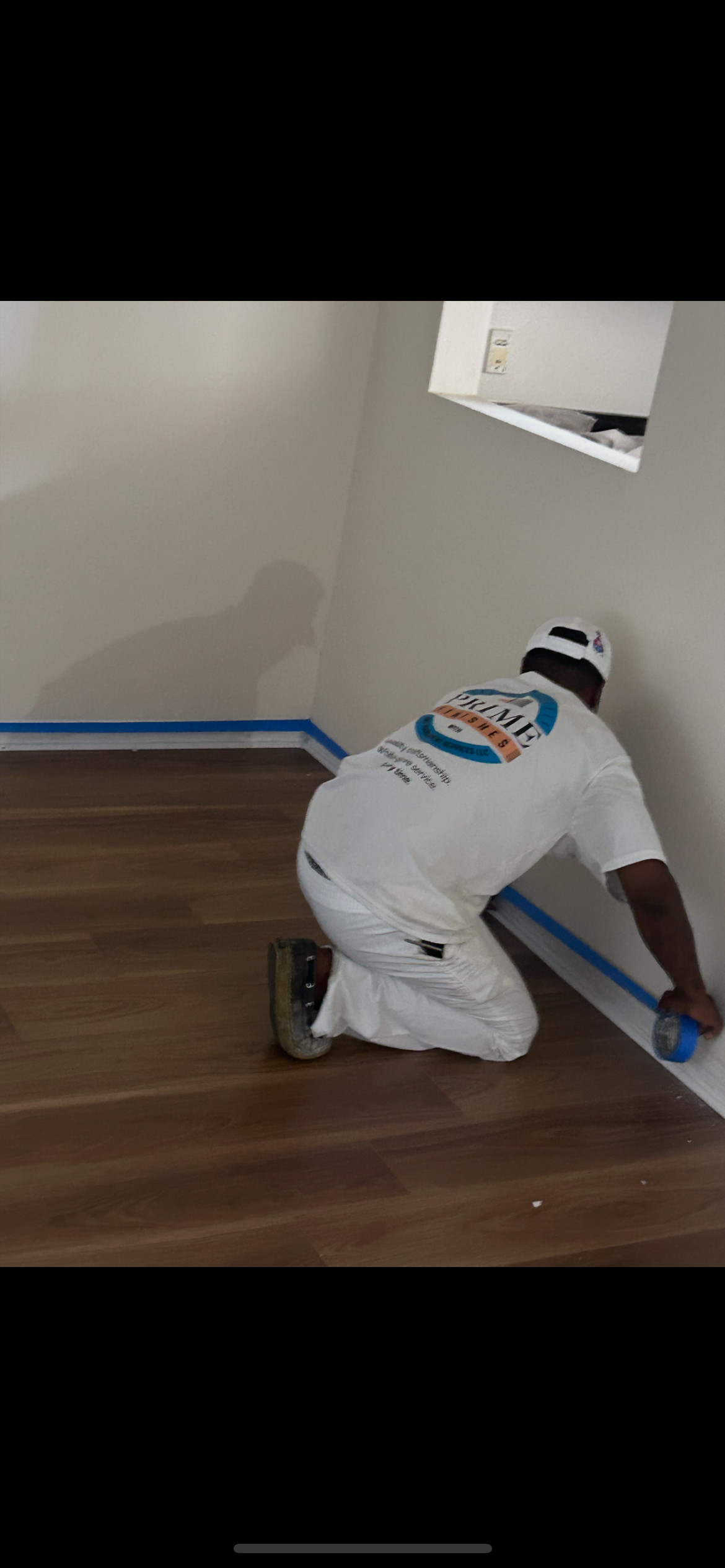 Painter in white overalls kneeling, applying blue tape along a wall and wooden floor.
