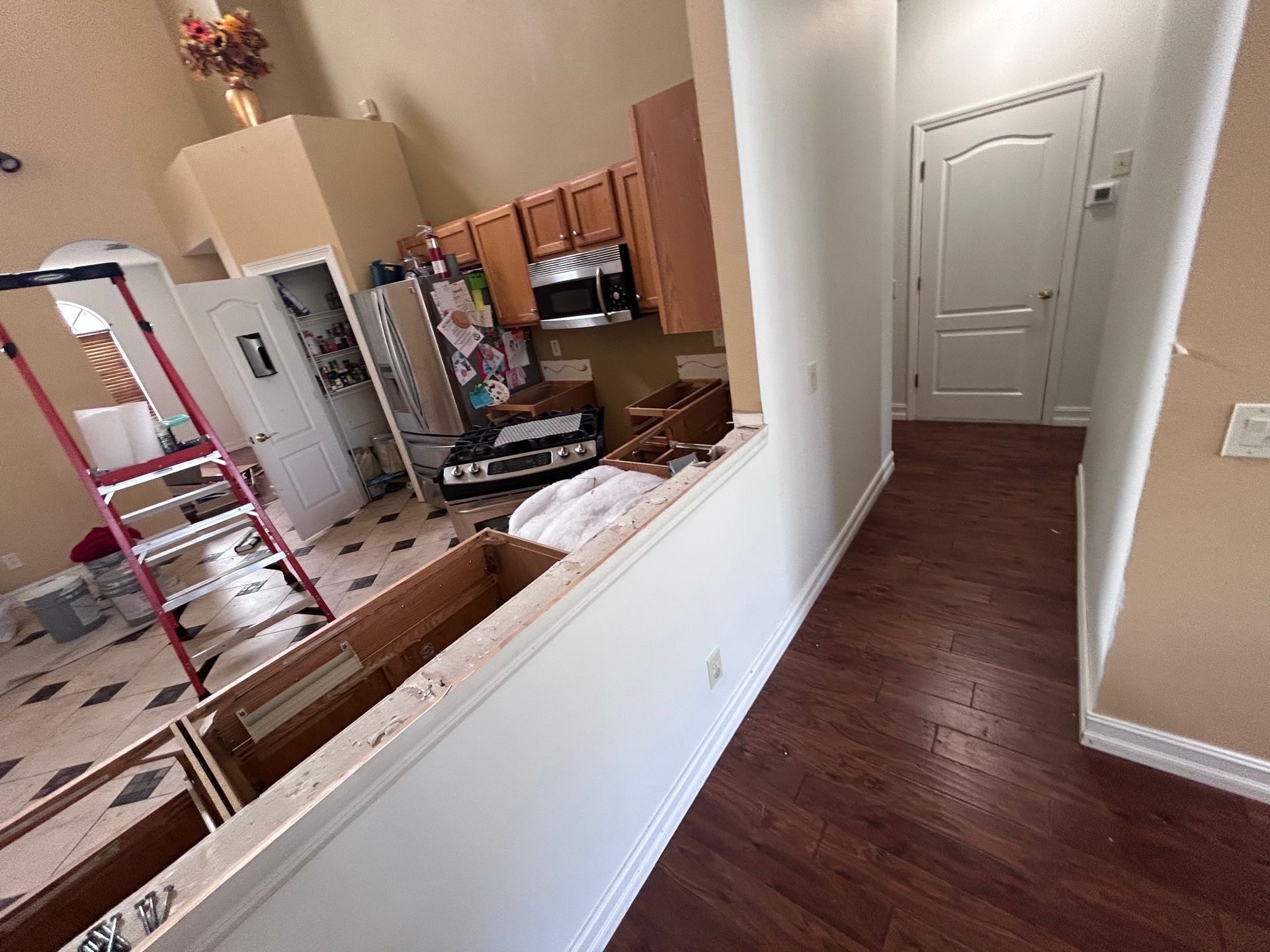 View of a kitchen and hallway; cabinets, appliances, and a red ladder are visible. Dark wood floors.