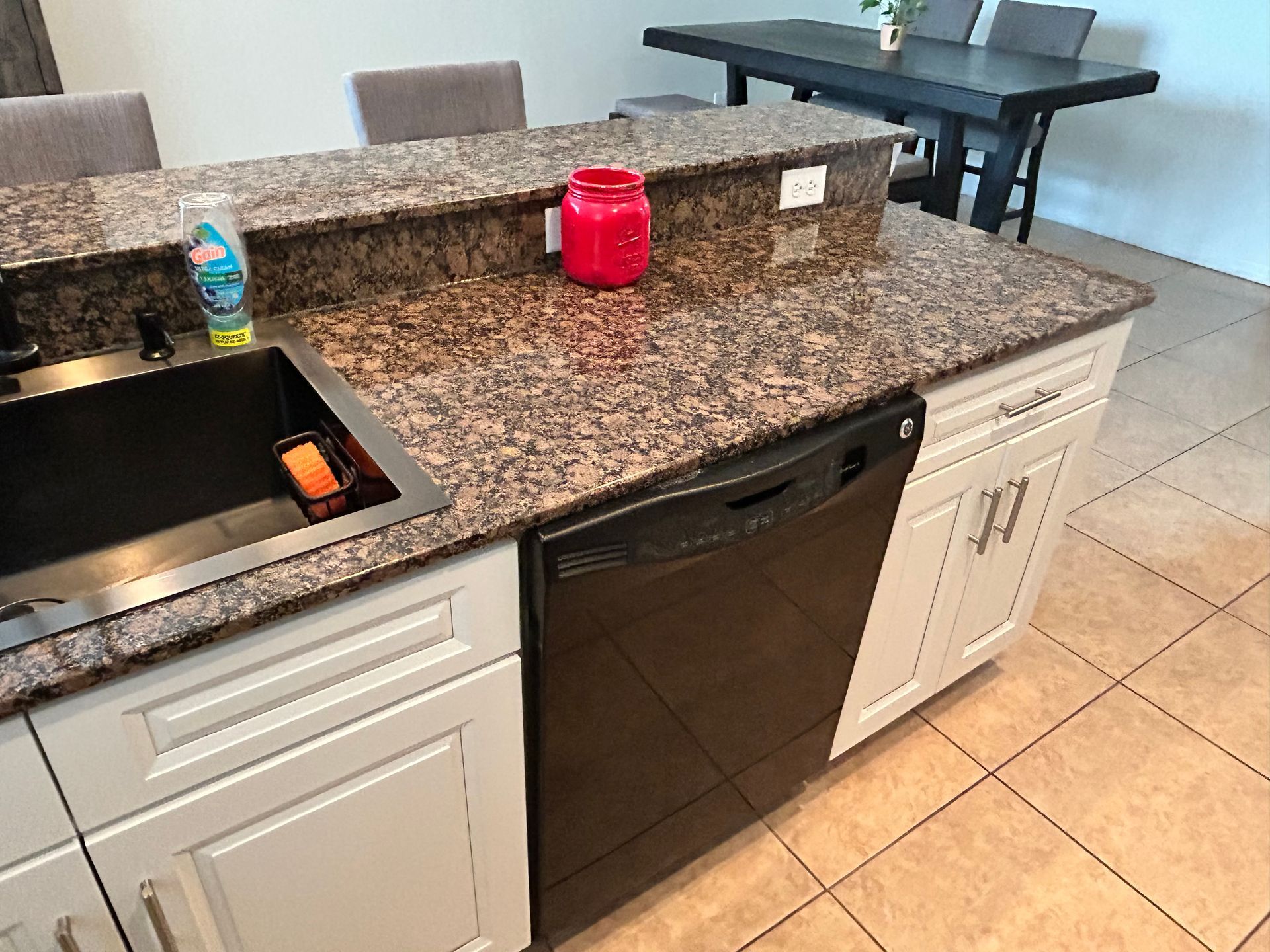 Kitchen island with granite countertop, black dishwasher, and white cabinets. Sink, red jar, and detergent visible.