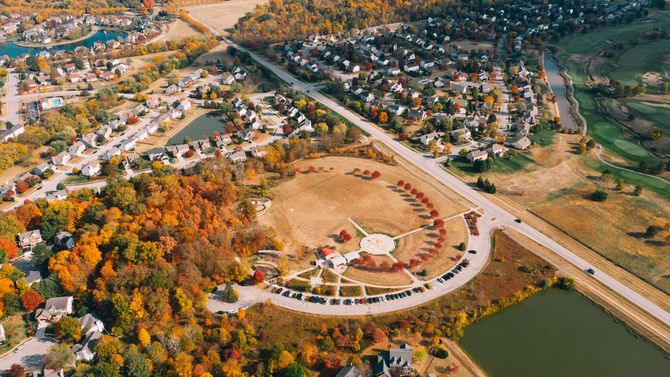 Aerial view of a circular park with autumn trees, surrounded by houses and roads beside a small lake