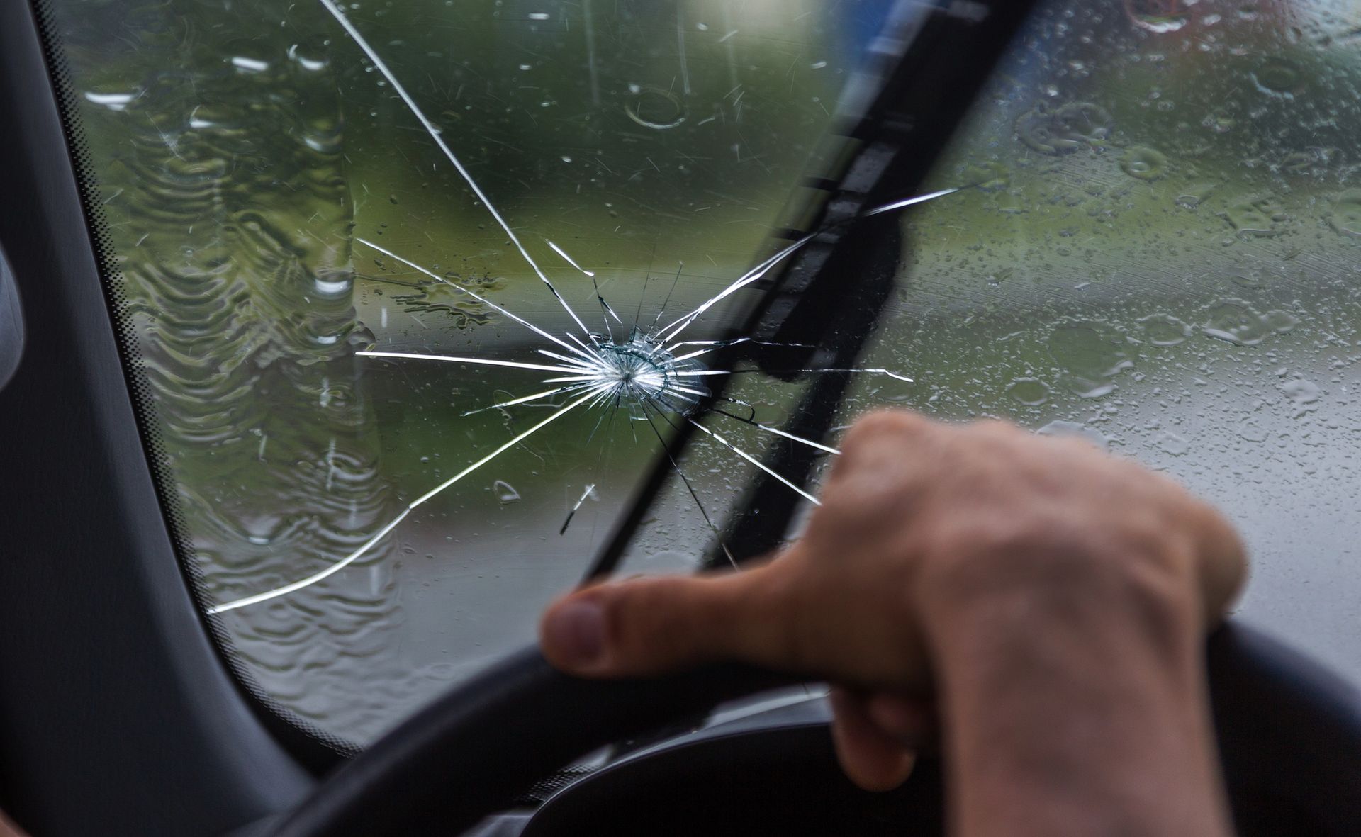 La mano di una persona stringe il volante davanti al parabrezza di un'auto, che presenta una grande crepa a forma di stella causata dall'impatto con un sasso.