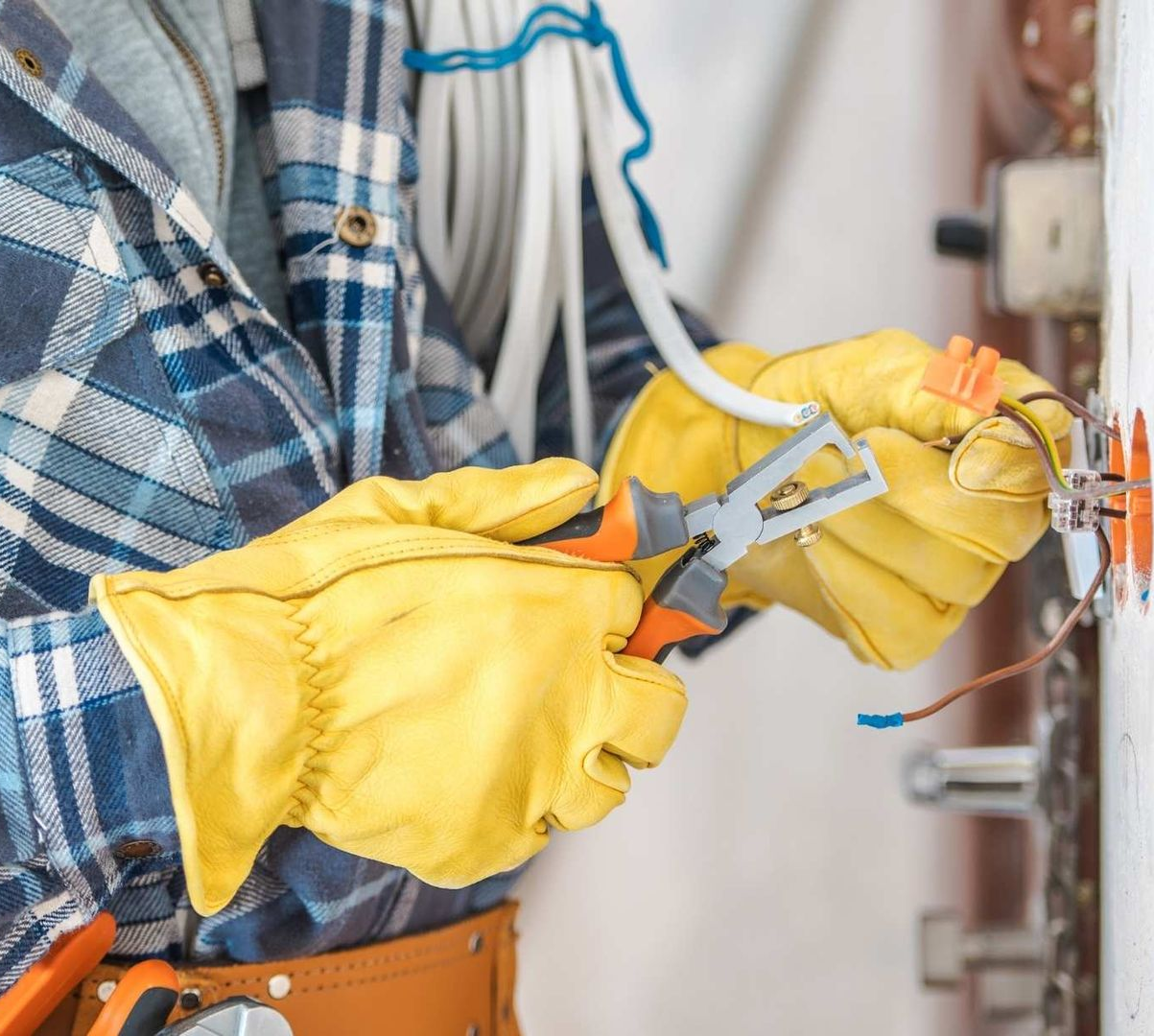Electrician wearing yellow gloves, working on wiring with pliers at a wall outlet.