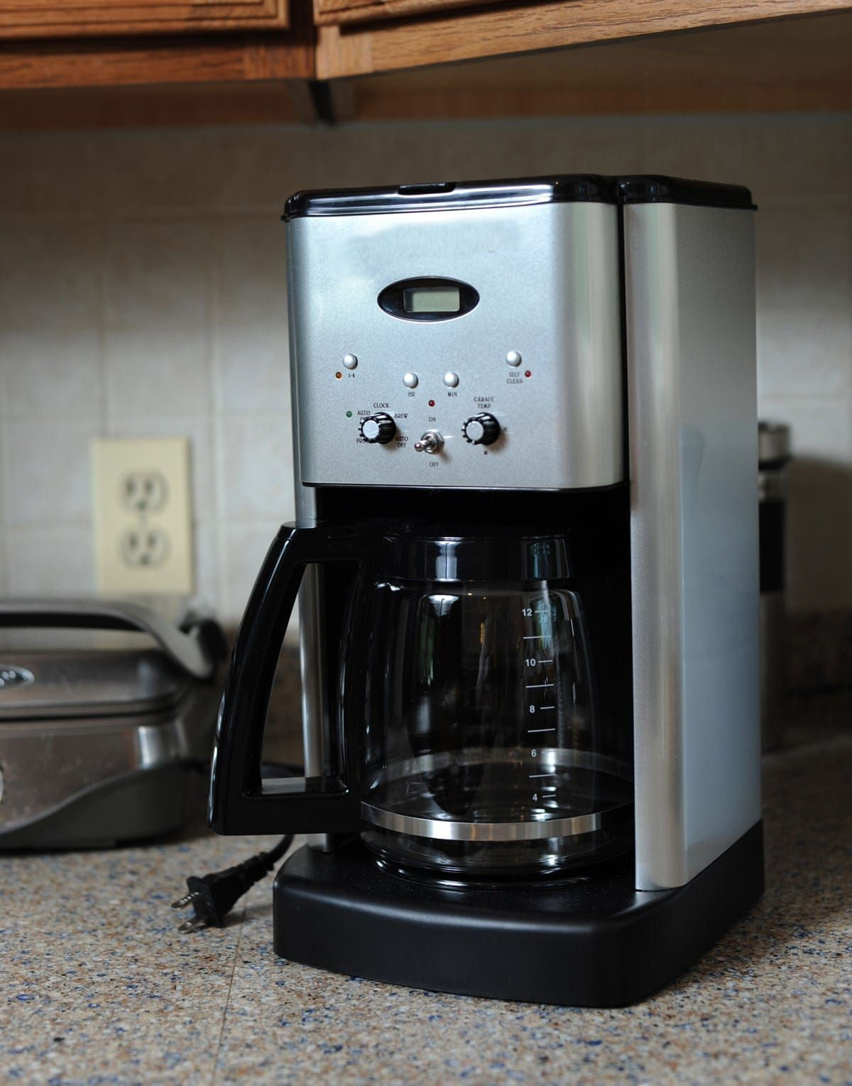 Silver coffee maker on a countertop, with a black carafe. Electrical outlet and toaster oven in background.