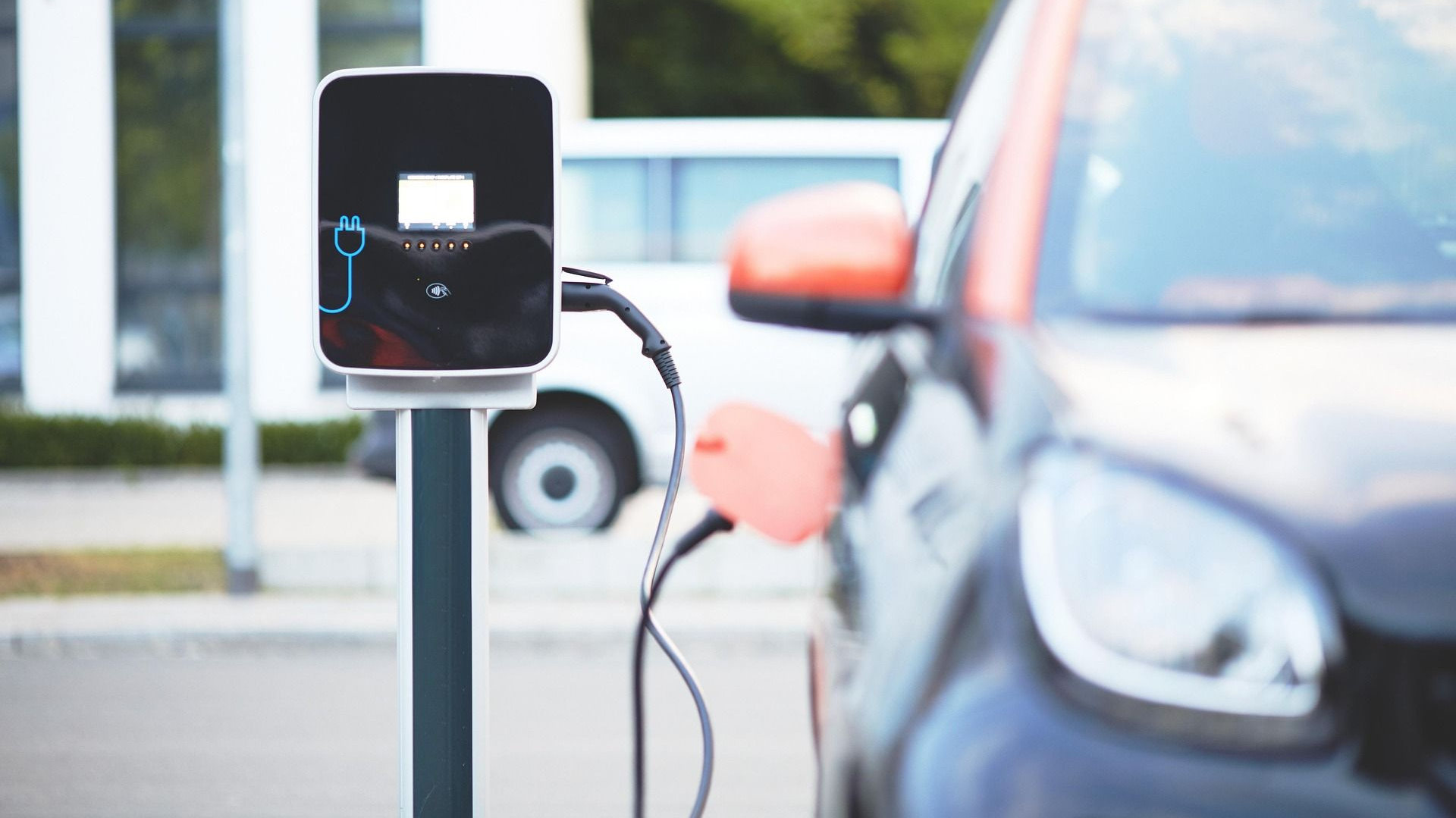 Electric car charging at a station with orange and black car, outdoors.