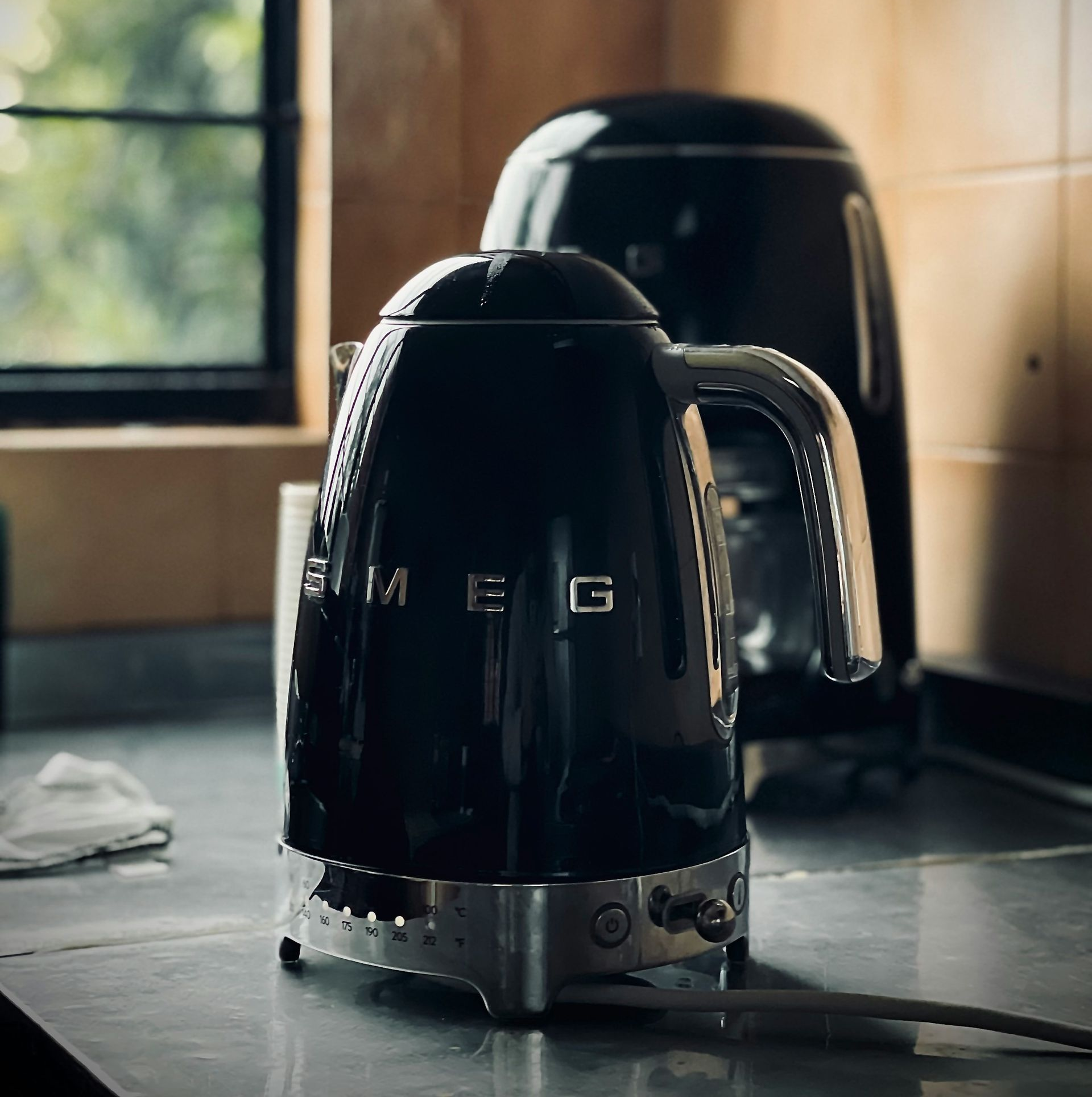 Black Smeg kettle with a matching toaster on a kitchen counter near a window.