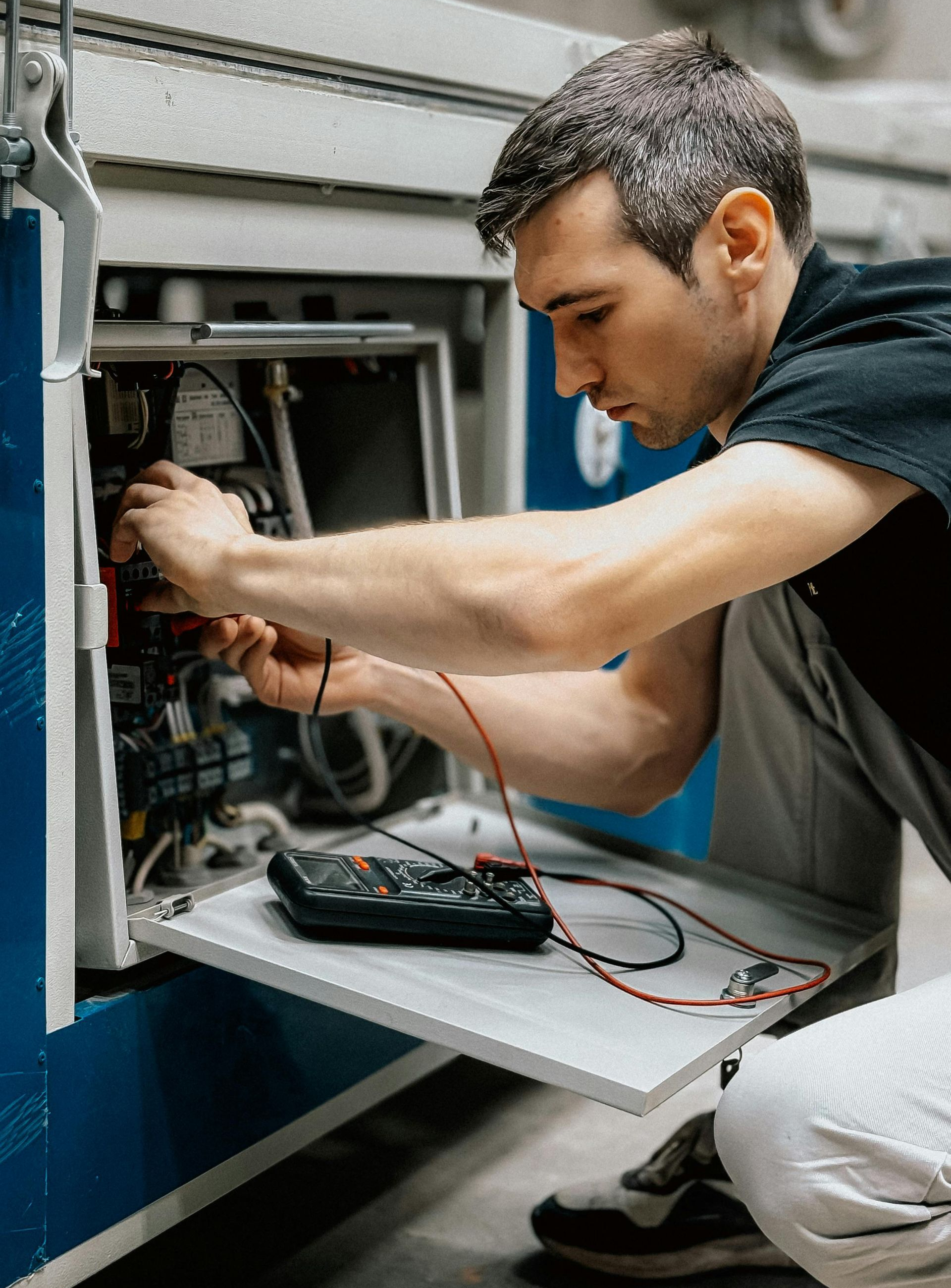 Man working on machinery, using a multimeter; interior setting.