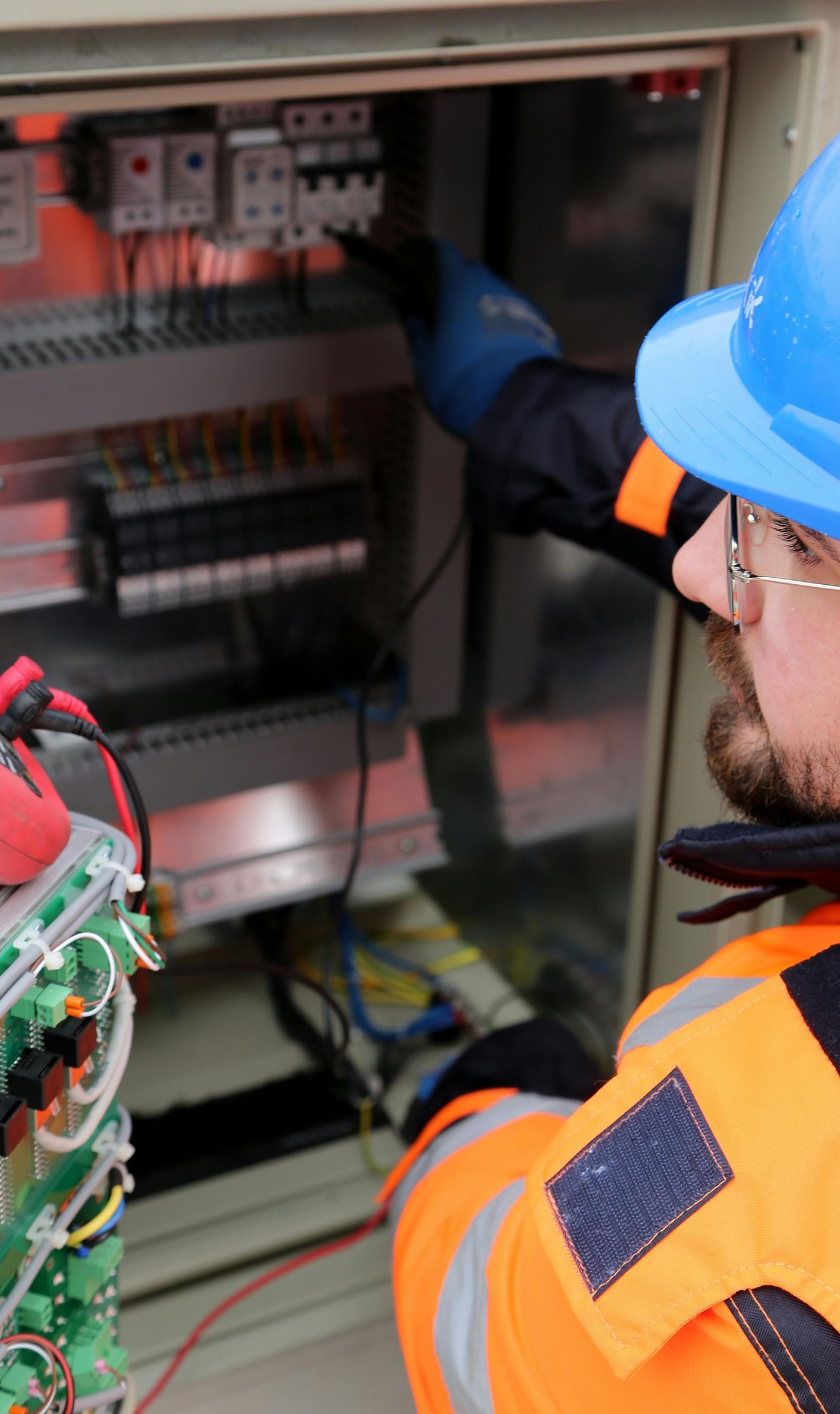 Electrician in blue hard hat tests electrical panel with multimeter. Orange safety vest and gloves.