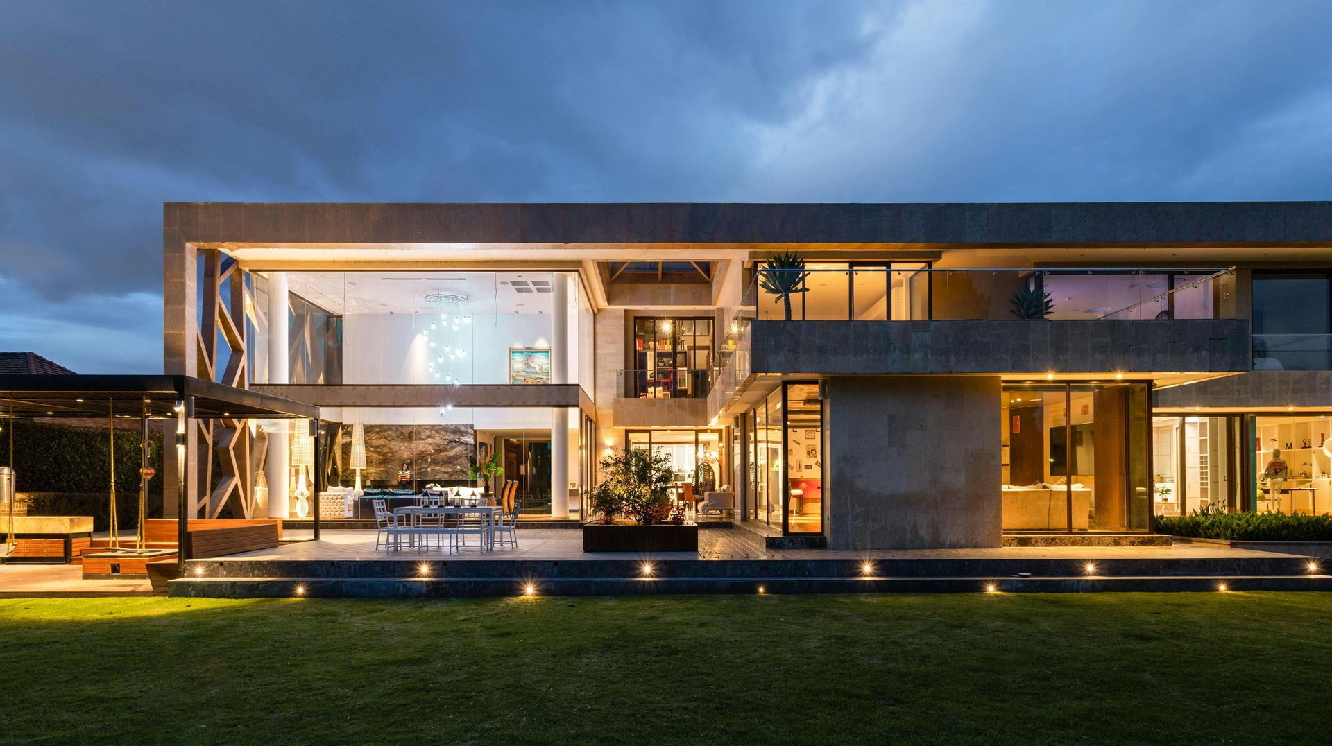 Modern two-story house at dusk with bright interior lights, a lawn, and cloudy sky.
