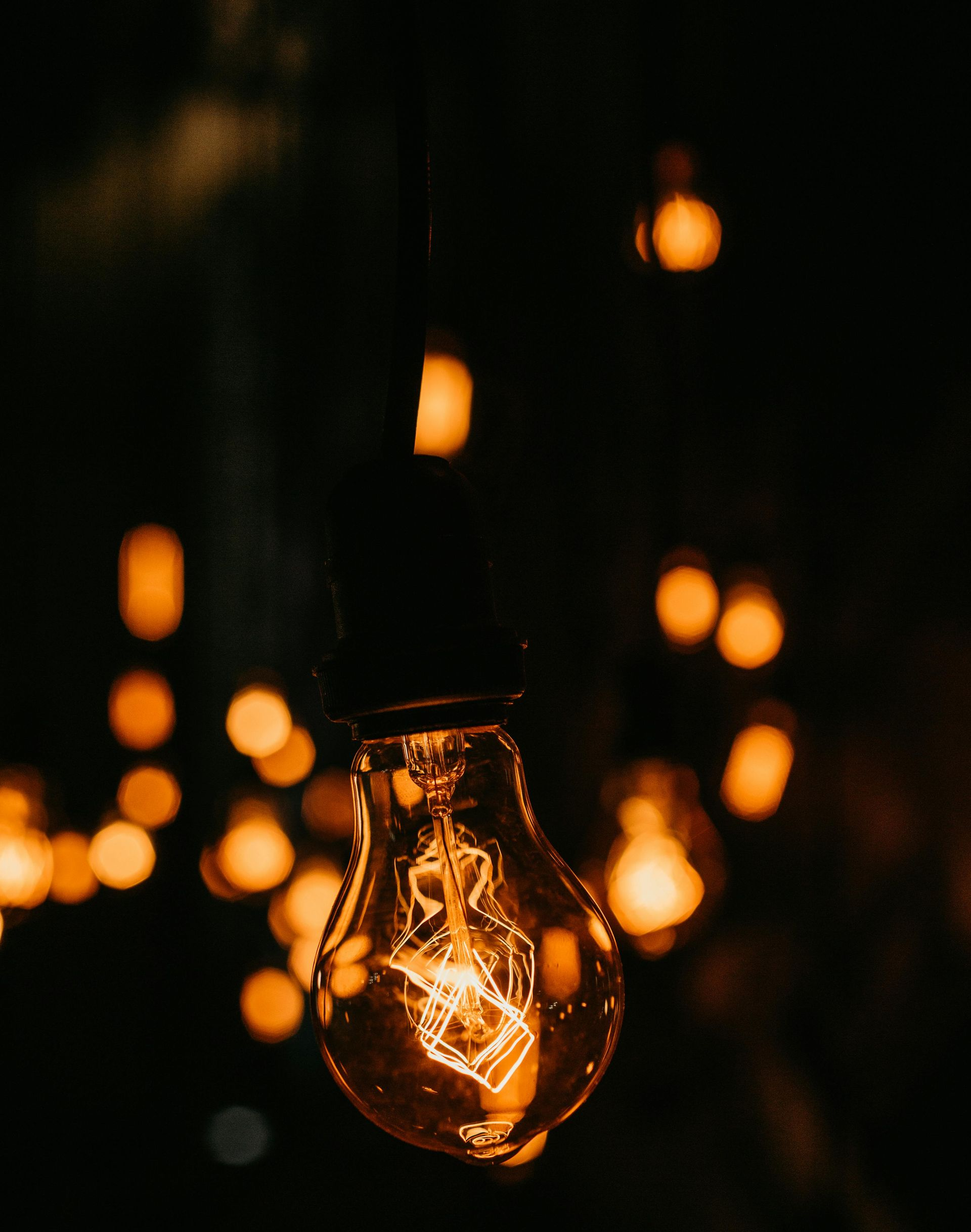 Glowing lightbulb with filament visible, surrounded by blurred orange lights in a dark setting.