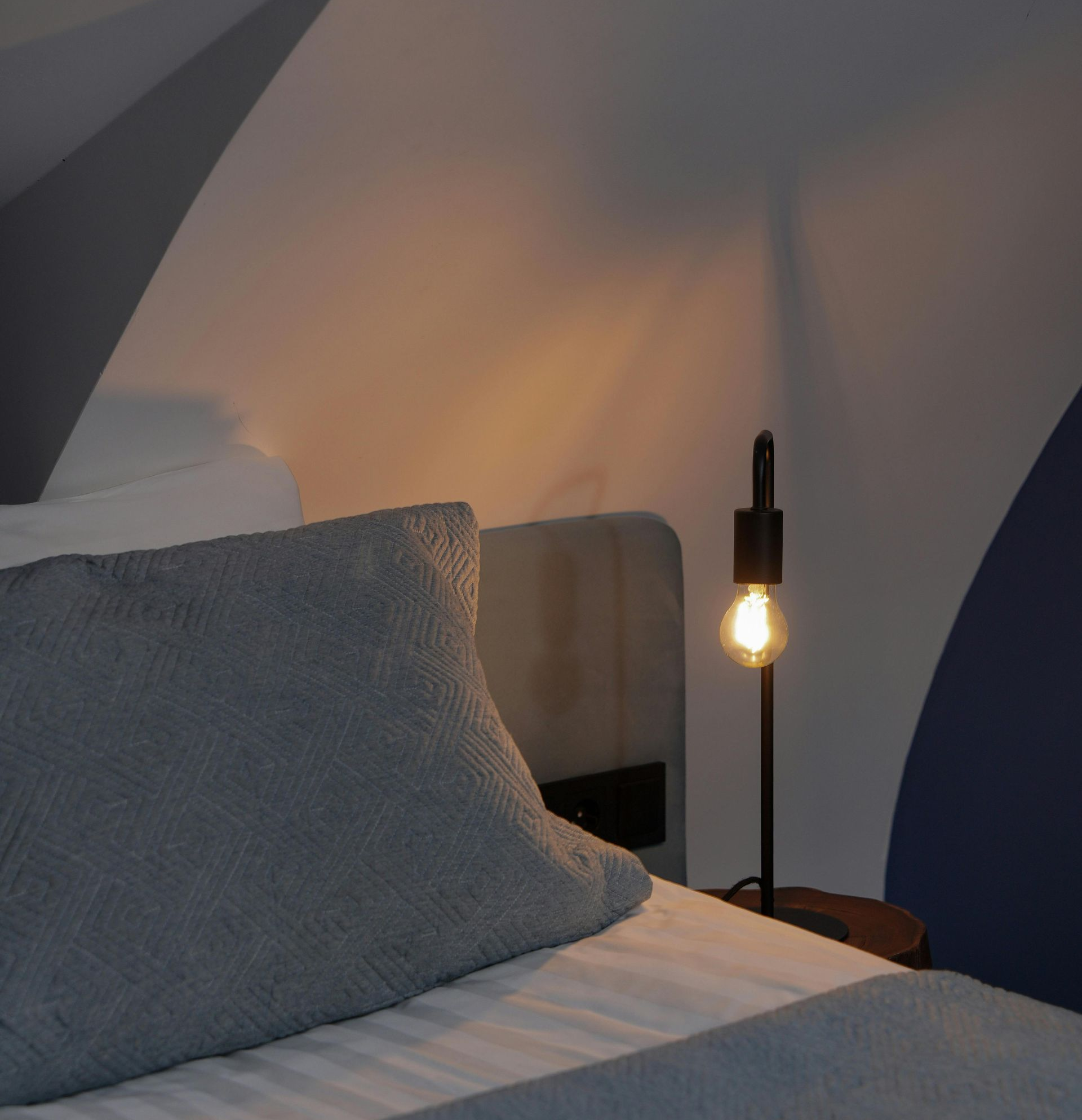 Bed with a gray pillow, lit lamp on a nightstand in a room with arched ceiling.