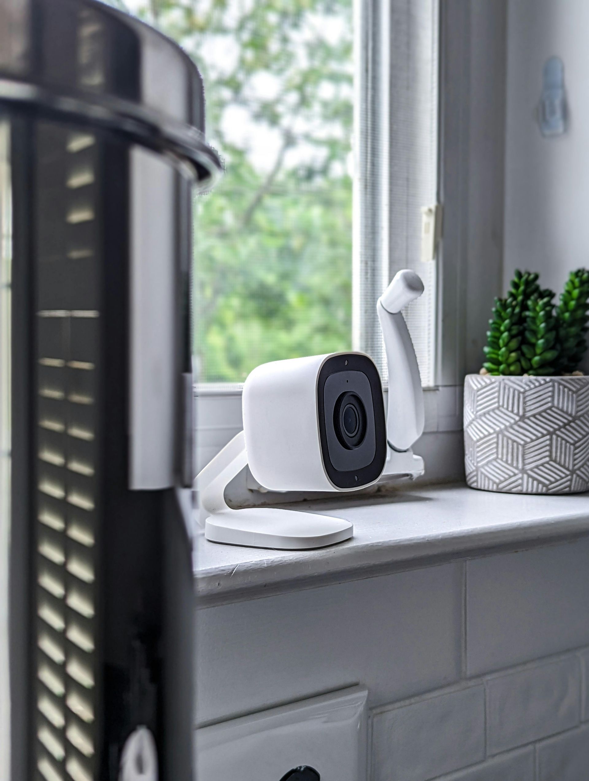 A white security camera sits on a windowsill next to a small potted plant, with a dark fan in the foreground.