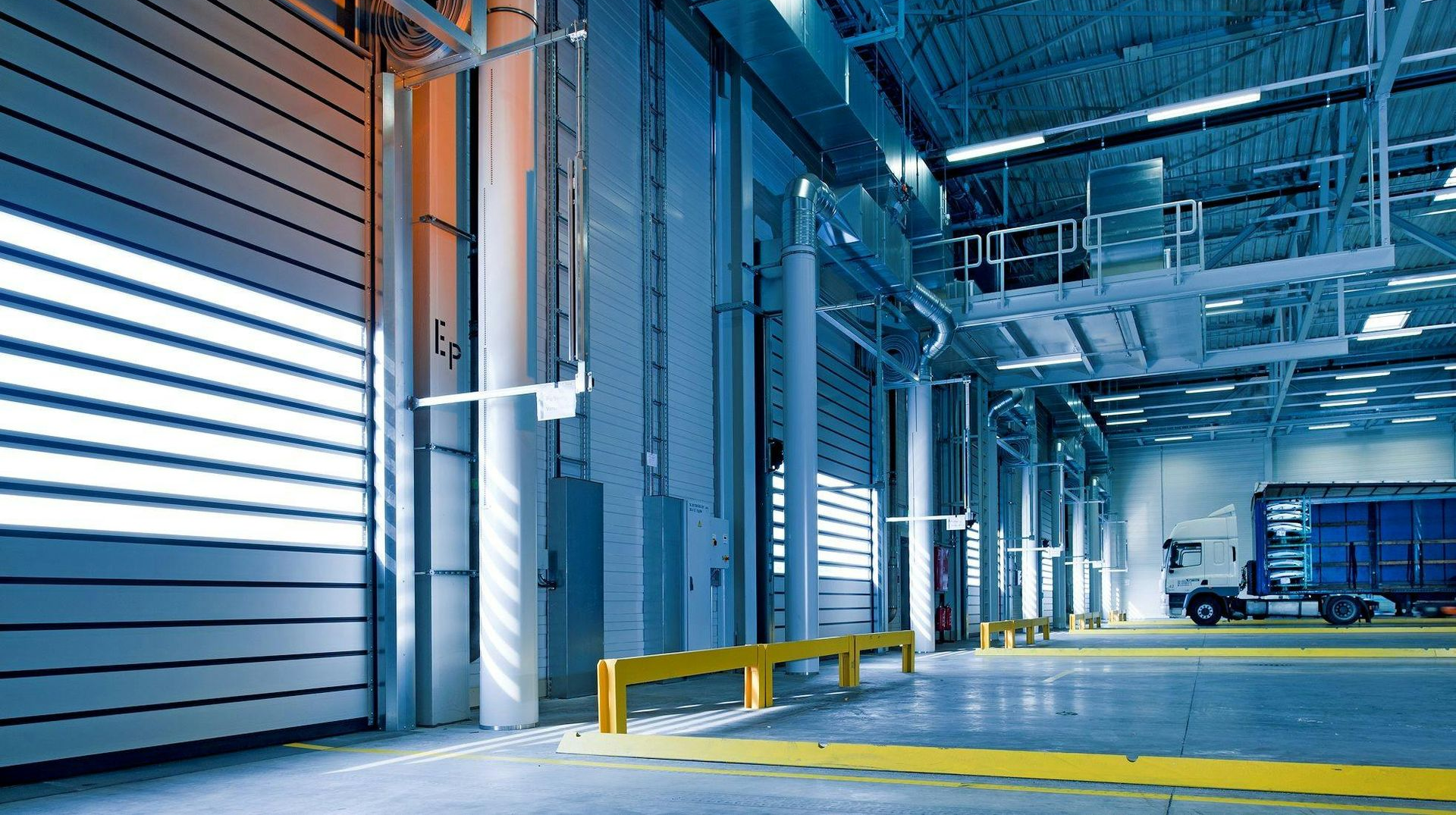 Loading dock interior with a truck, roll-up door, and yellow safety barriers.