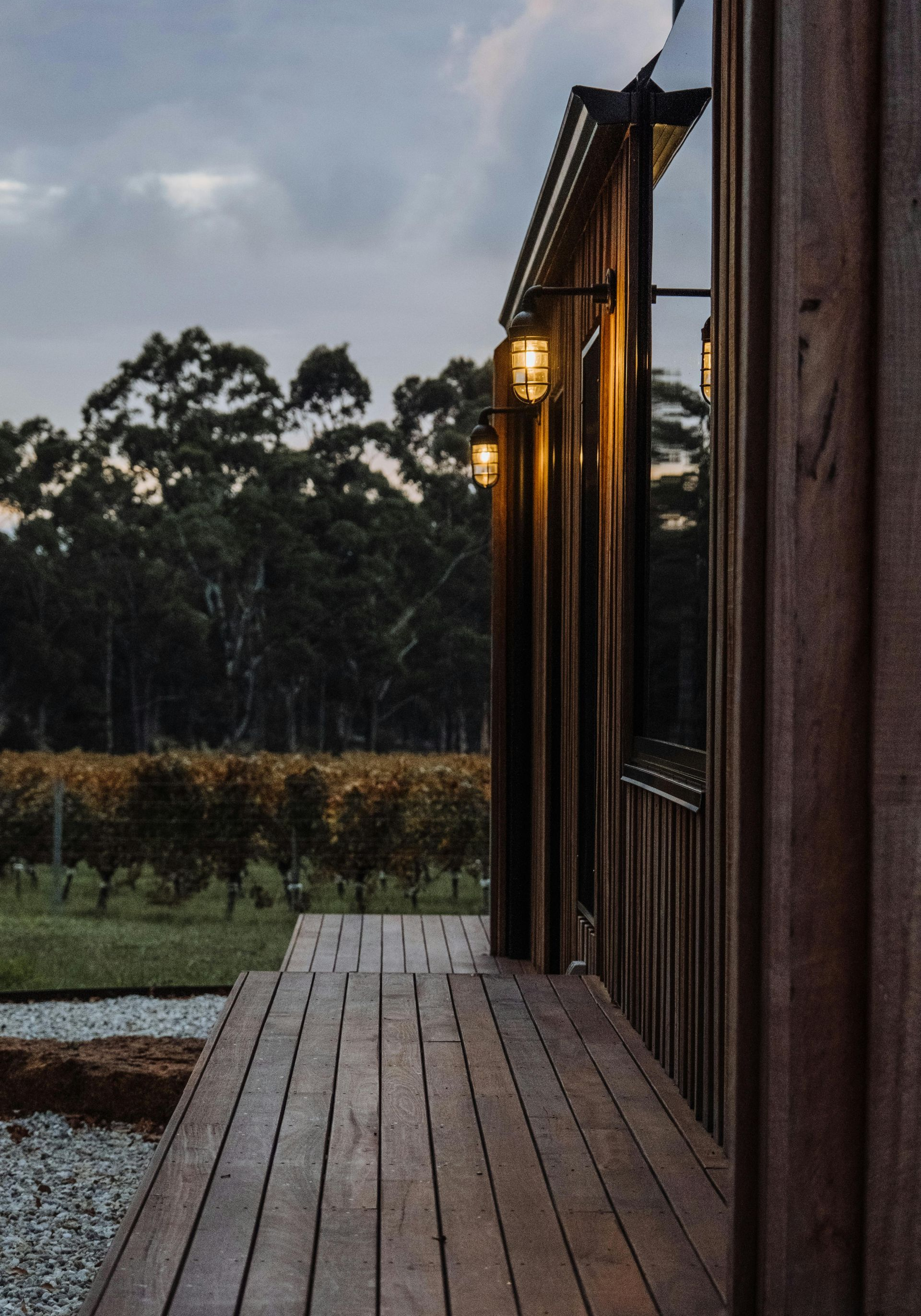 Wooden cabin exterior with deck, wall lights, overlooking a vineyard at dusk.