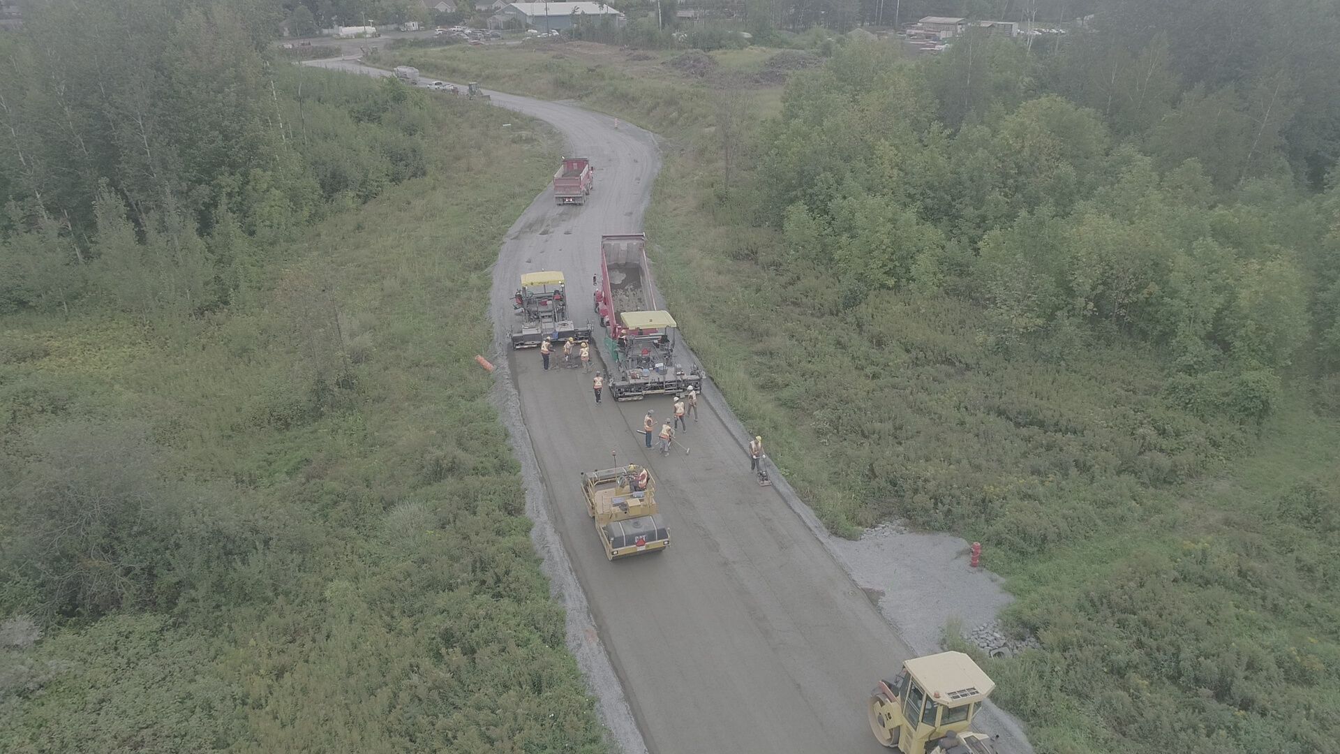 Des engins de chantier lourds travaillent sur un chemin de terre sinueux traversant une végétation dense.