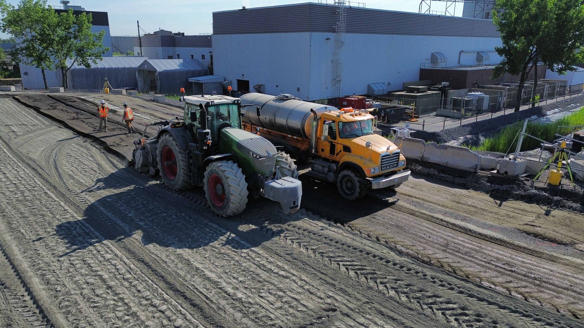 Des engins de chantier effectuent des travaux de pavage sur une route longeant un grand bâtiment, avec des ouvriers et des cônes de signalisation à proximité.