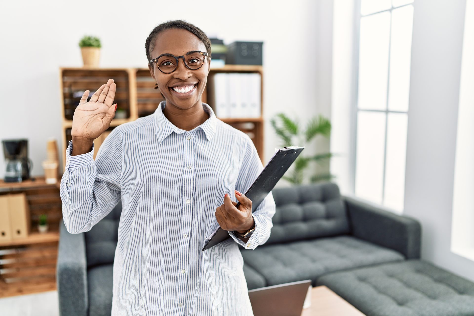 Woman in glasses, holding clipboard, waving at the camera in a modern office setting.