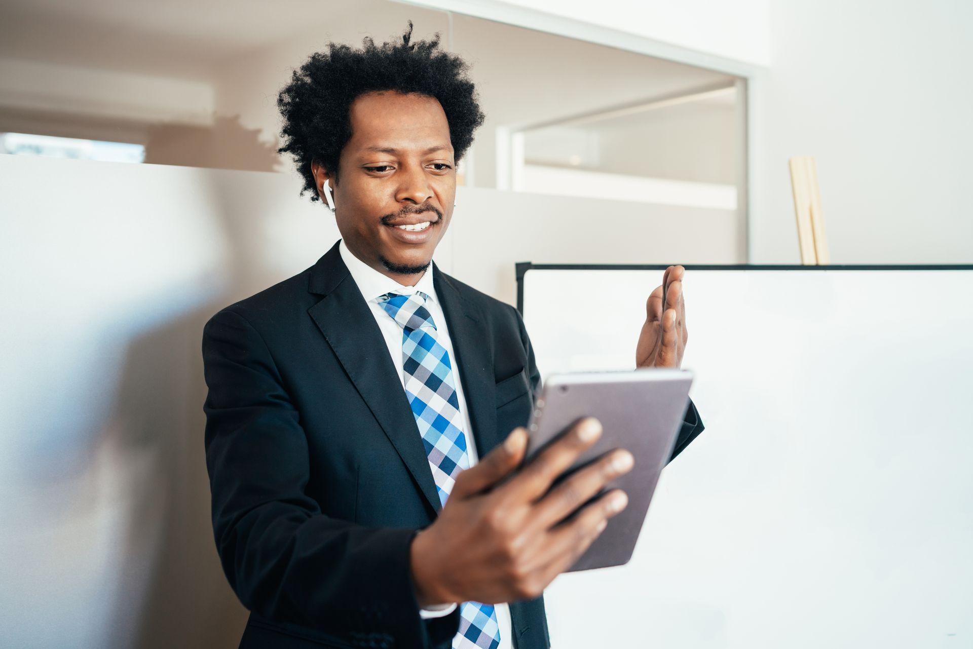 Man in suit holding a tablet, gesturing during a video call in front of a whiteboard.