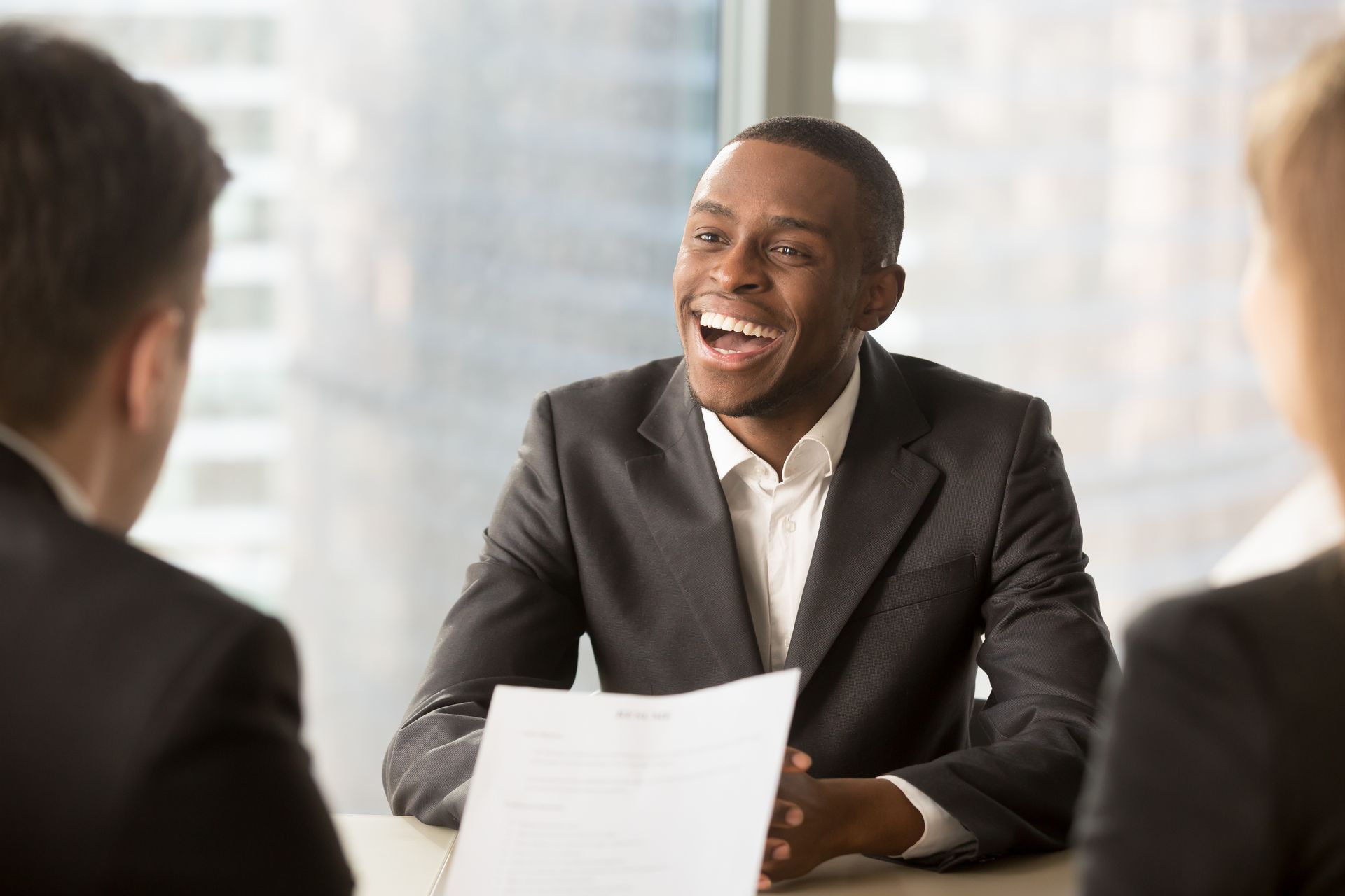 Smiling man in a suit at a table, facing two people and holding papers in a bright office.