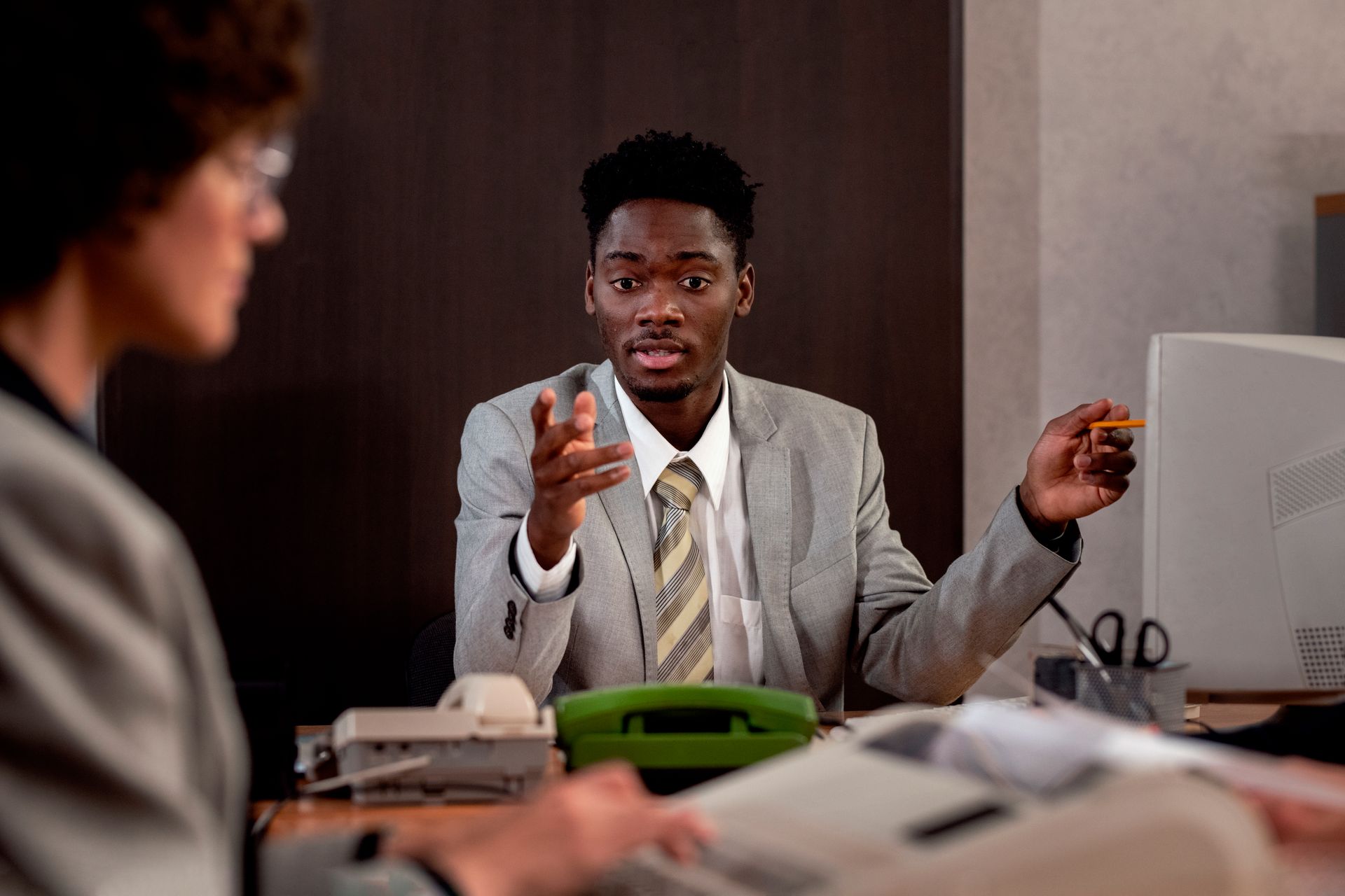Man in suit gesturing while speaking at a desk, facing another person out of view. Office setting.